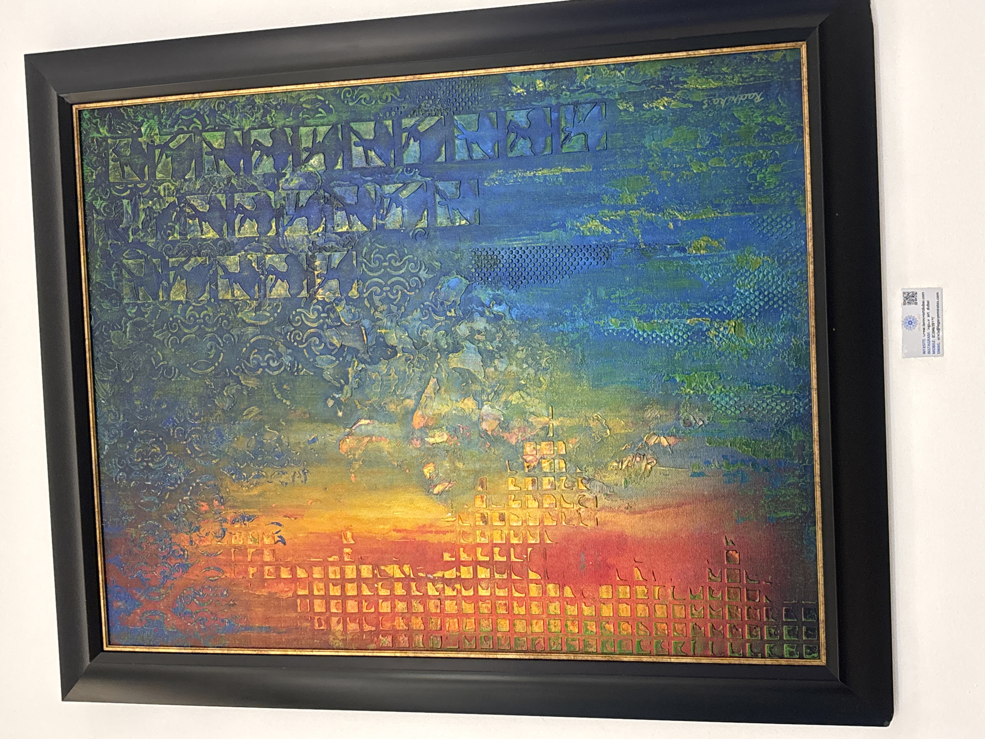 The image shows a framed abstract painting displayed on a white wall. The painting features a vibrant gradient of colors transitioning from red and orange on the left to yellow, green, and blue on the right. The surface of the painting has a textured appearance with various patterns, including a grid of squares on the left, intricate floral designs, and what appear to be stylized figures or shapes in a grid on the right. A signature, "Radhika," is visible in the lower right quadrant of the painting.
Below the painting, a small white label with a QR code and text is attached to the wall. The text on the label reads: "WEBSITE: www.foreverartdxb.com / INSTAGRAM: @foreverartdxb / DUBAI / MOBILE: 0504467917 / EMAIL: art@foreverartdxb.com". This label indicates that the artwork is likely displayed in Dubai, United Arab Emirates, possibly at a gallery or exhibition, and provides contact information for the artist or gallery. The city is identified as "Dubai" in the text on the label, and the context suggests that "Ff" in the prompt is likely a typo or refers to Dubai.
