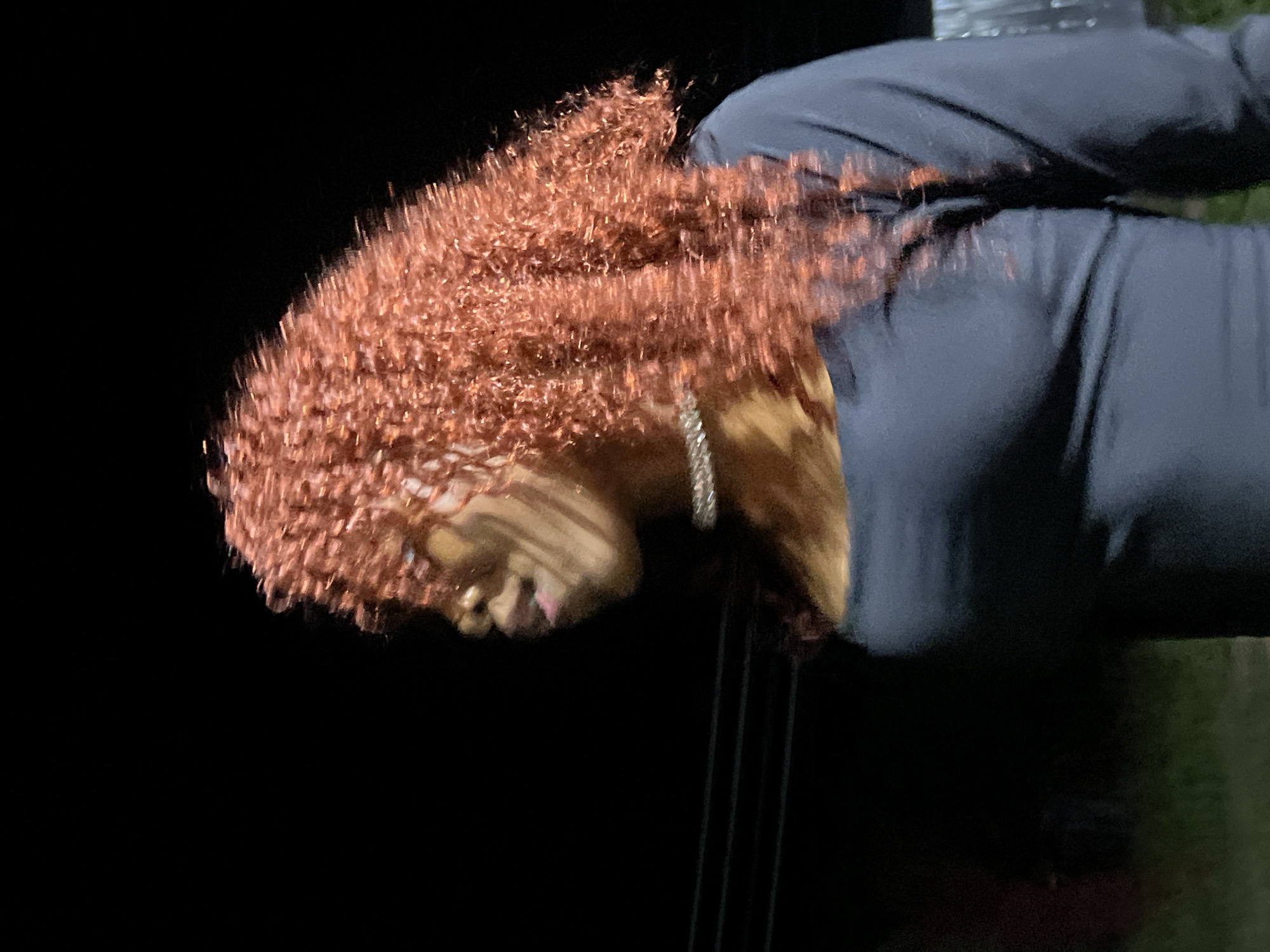 The image shows a side profile of a Black woman with curly, reddish-brown hair, wearing a dark-colored, long-sleeved shirt and a sparkly necklace. She is standing outdoors at night, with a dark background that suggests the absence of artificial light sources. The image is somewhat blurry, indicating motion blur or low light conditions. There are indistinct horizontal lines visible in the background, possibly power lines or branches. The woman's face is partially visible, with her mouth slightly open. The environment appears to be a natural or semi-natural setting, as suggested by the faint hint of green at the bottom of the frame, which could be grass or foliage. The overall lighting is dim, with some highlights on the woman's hair and necklace. The location is specified as Durumi, Nigeria.