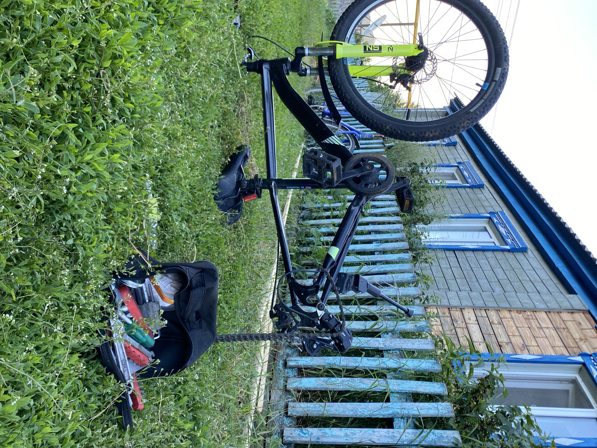 The image shows a bicycle leaning against a wooden fence in front of a house. The bicycle is black with a neon yellow front fork and a black seat. It has a black bag attached to the rear, filled with various tools, including red and green items that appear to be pliers and wrenches. The bicycle's chain and derailleur are visible. The fence is made of weathered blue and natural wood planks, with some greenery growing through it. Behind the fence is the facade of a wooden house with blue trim and windows. The house appears to be in a rural setting, consistent with the provided location context of Maloarslangulovo, Russia. The lighting suggests it is daytime, possibly late afternoon or early evening, with the sun casting shadows. The overall scene evokes a sense of a rural homestead, perhaps a place where repairs are done outdoors. Text visible on the bicycle's fork reads "NS N".