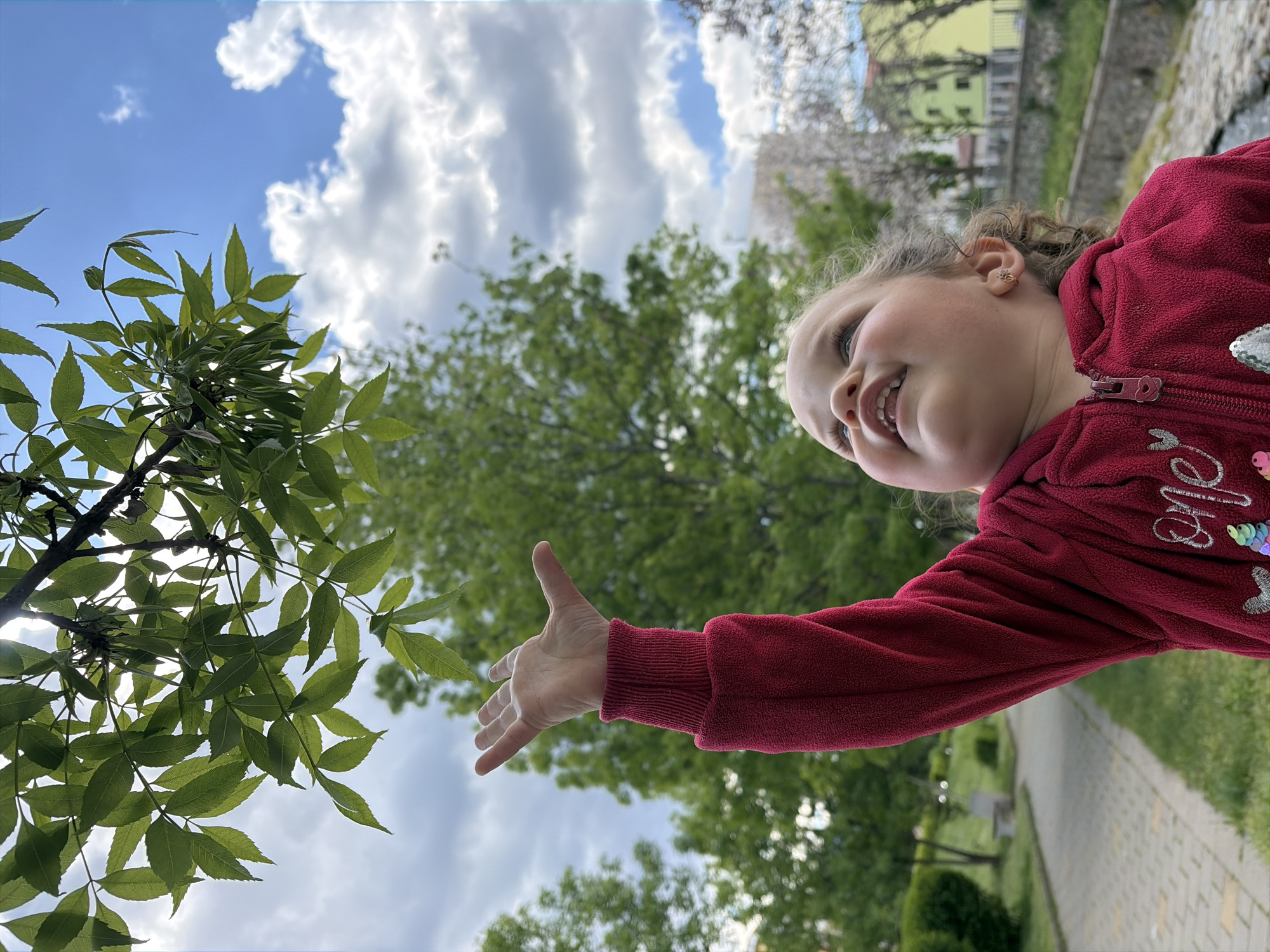 A young girl is reaching towards a tree with green leaves against a sky with scattered clouds. The girl is wearing a red zip-up jacket. The scene is outdoors, with a paved pathway and greenery visible in the background. A building is also partially visible in the background. The location is Unknown, Unknown.