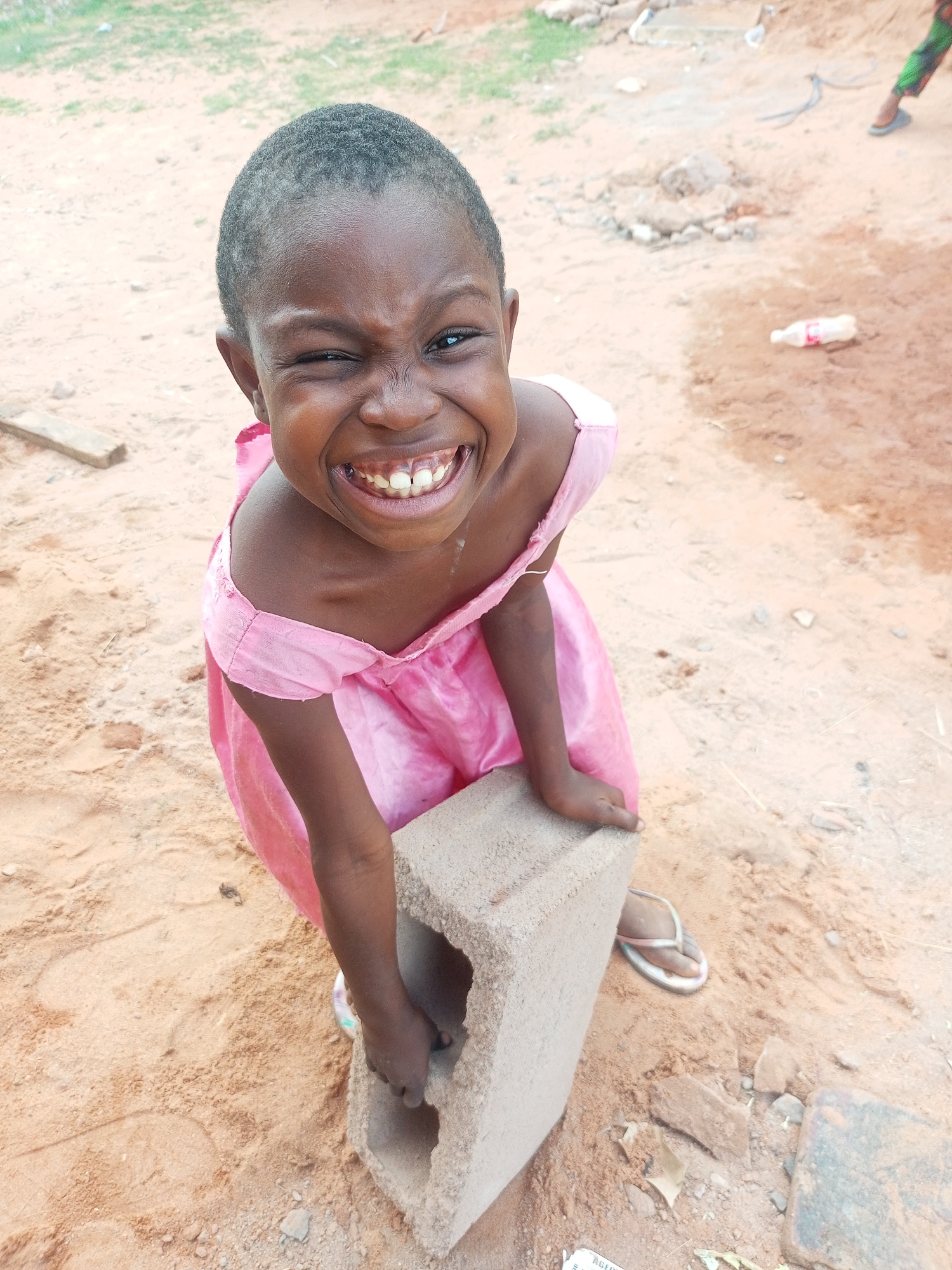 This outdoor portrait captures a young girl in Tempure, Nigeria, with a beaming, joyful expression. She is the central subject, looking directly at the viewer with a wide smile that crinkles her eyes, giving the appearance of a wink. Her short, dark, curly hair frames her face.
The girl wears a bright pink, off-the-shoulder dress, which appears somewhat worn. She is positioned next to a large, light grey hollow concrete block, gripping it with both hands. Her left foot, wearing a light-colored flip-flop, is visible on the sandy ground.
The setting is an undeveloped, sandy area, possibly a construction site or a rural village environment. The ground is predominantly light brown sand with scattered small stones, debris, and patches of sparse greenery in the background. Other objects faintly visible in the background include a discarded plastic bottle and what appears to be a piece of wood. The lighting suggests it is daytime, likely with bright, natural light.
The dominant emotion conveyed by the image is pure happiness and playfulness, emanating from the girl's expressive face. Her engagement with the camera is direct and lively.
Near her foot, on the ground, there is a piece of paper or cardboard where the partially visible text "AGENT" can be faintly discerned.