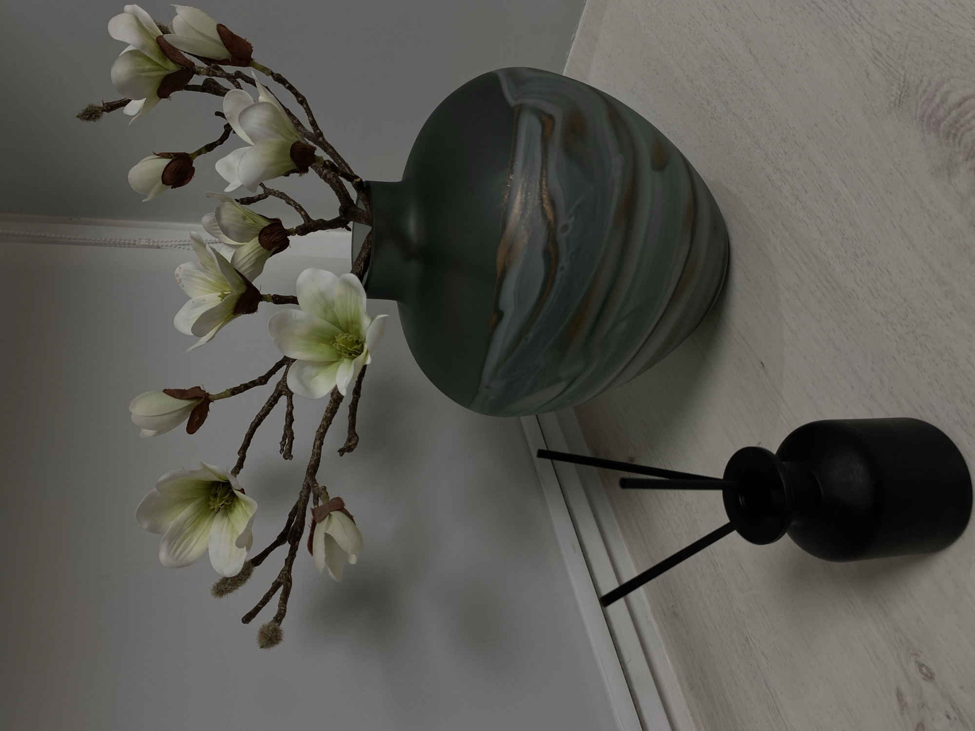 This media file features a still life arrangement of decorative items on a light-colored, wood-grained surface. The primary objects are a vase of artificial white flowers and a black reed diffuser.
The vase is a substantial, ovate piece with a muted, grayish-green base color. It is adorned with swirling patterns of white and gold, creating a marbled or aged effect. The vase is positioned on its side, leaning against a white vertical element, possibly a wall or partition.
Arranged in the vase are several artificial magnolia blossoms on brown stems. The flowers are white with hints of pale green in their centers. Some buds are also visible, along with fuzzy brown tips on the stems, adding a textural contrast. The flowers cascade downwards and to the left of the vase.
To the right of the vase, and slightly lower, is a matte black reed diffuser. It has a rounded body and a narrow neck topped with a black disc, from which two black reeds extend upwards and to the left, creating a balanced visual composition with the flowers.
The background is a plain, light gray surface, providing a neutral backdrop that emphasizes the objects. The lighting appears to be artificial, casting soft shadows and illuminating the scene from above and to the right. The overall impression is one of quiet, decorative elegance, suitable for interior styling or a staged product shot. There is no visible text in the image. The scene is indoors, and there are no indicators of specific time of day or weather conditions.
