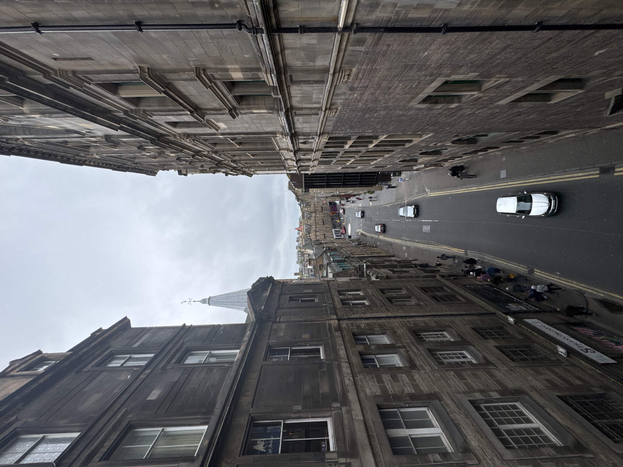 The image shows a view from above, looking down a narrow, wet street lined with old stone buildings in the city of Edinburgh, United Kingdom. The architecture is predominantly old, with many windows and some decorative stonework visible on the facades. The street is paved and has double yellow lines marking the center. Several cars are driving or parked on the street, and a few pedestrians are walking along the sidewalks. The sky is overcast and gray, contributing to a somber atmosphere. In the background, more buildings with similar architectural styles are visible, along with a distinctive church spire. The scene appears to be a typical urban street in an older part of the city.