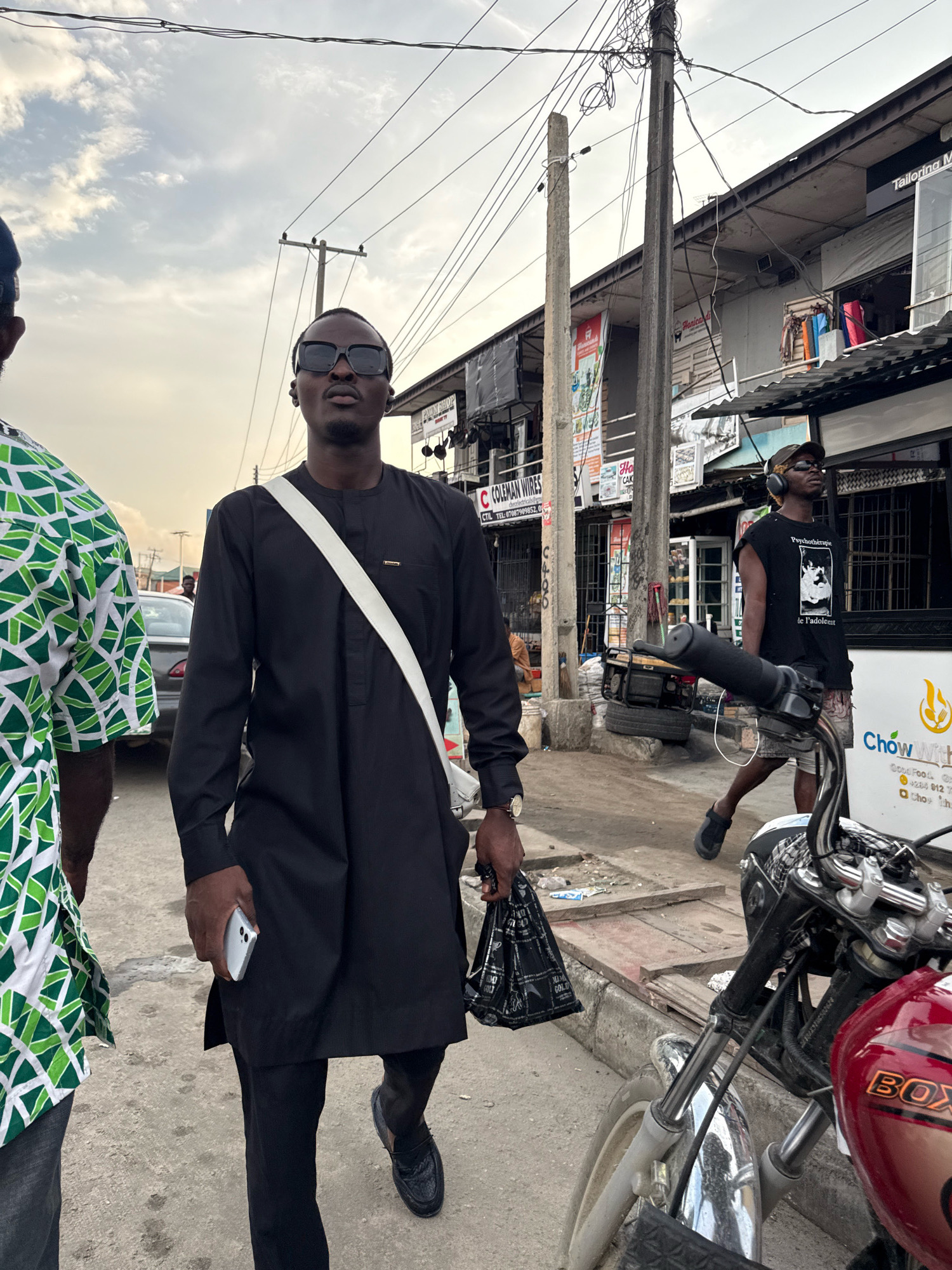 The image captures a street scene in Ado, Nigeria, under an overcast sky. In the foreground, a man wearing sunglasses and a black traditional outfit walks towards the camera, a white strap slung across his chest and a black plastic bag in his left hand. He is holding a white smartphone in his right hand. To his left, another person is partially visible, wearing a green and white patterned shirt. In the background, there are buildings with signs, including one for "Tailoring Master" and another with "Coleman Wires." A motorcycle is parked in the lower right portion of the frame. The street appears to be paved, with a curb and some debris visible. Utility poles with numerous wires are prominent features of the urban landscape.