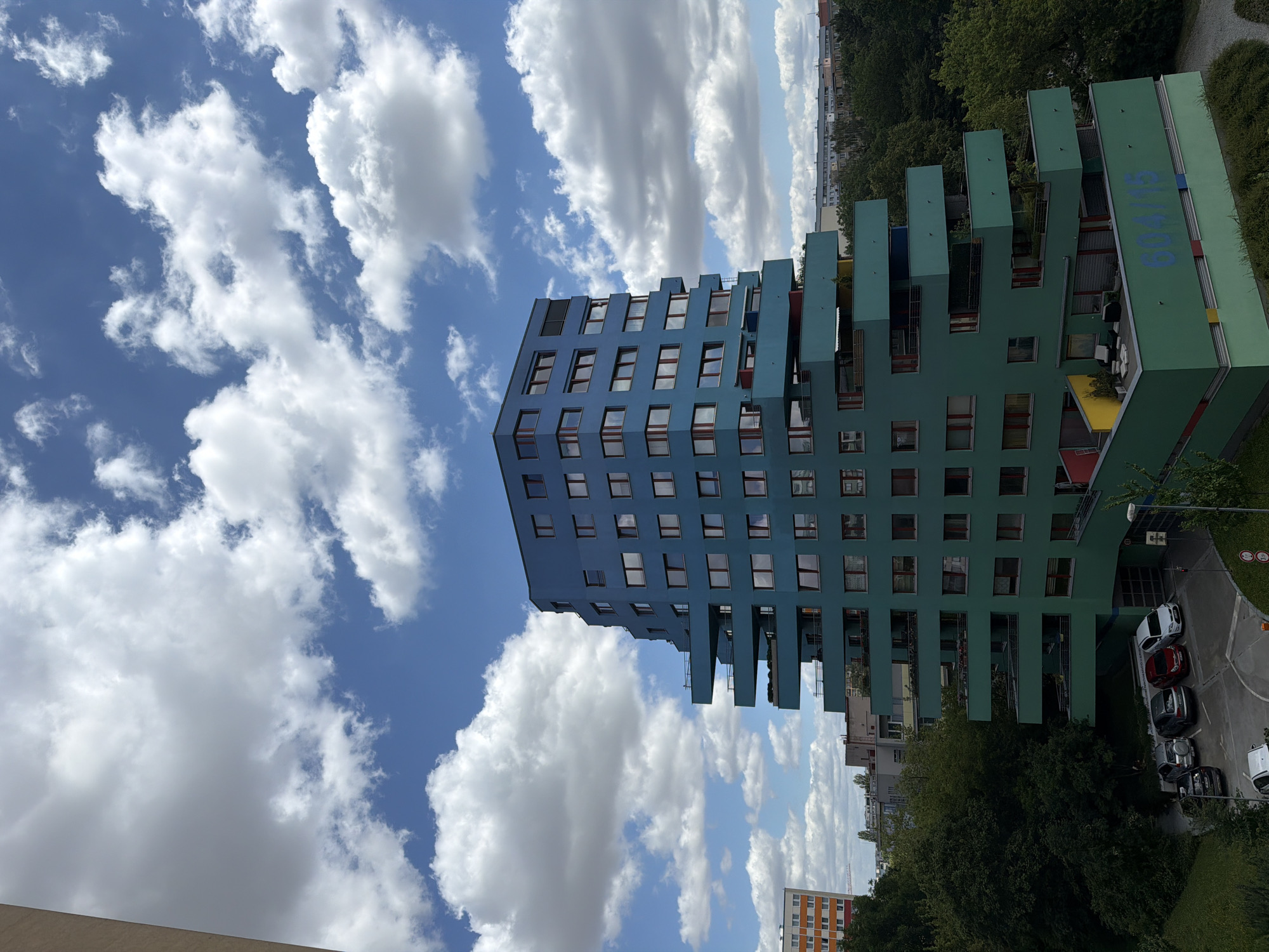 A wide-angle, eye-level shot captures a modern, multi-story building with a distinctive tiered design in the city of Prague, Czechia. The building, primarily teal blue and green, features numerous balconies that jut out at various angles. The sky above is a clear blue, dotted with fluffy white cumulus clouds. In the foreground, several parked cars are visible in a paved lot, with lush green trees and other buildings forming the surrounding urban landscape. The facade of the building prominently displays the numbers "604/15" in blue lettering.