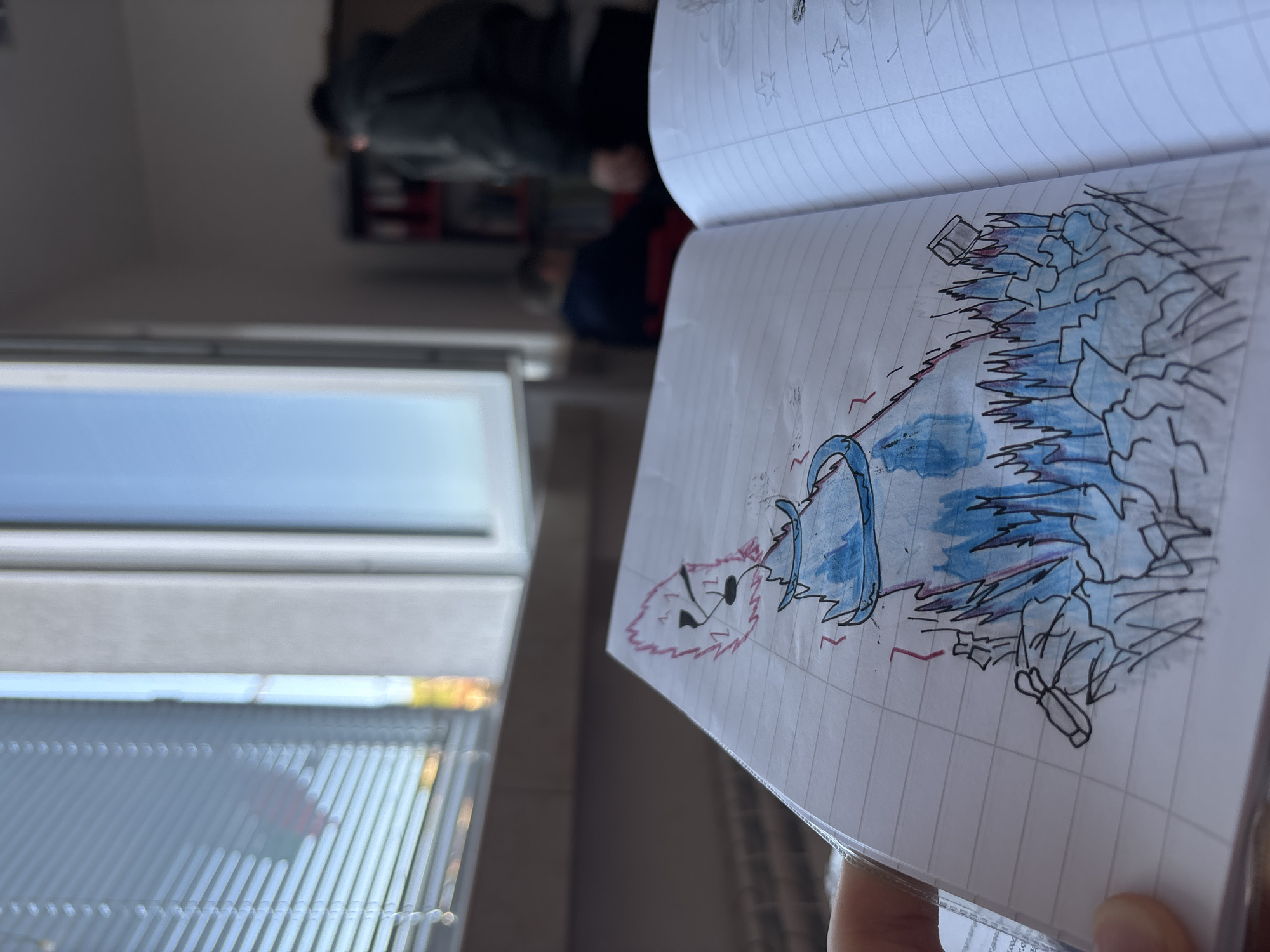 The image, rotated counter-clockwise, features a person's hand holding an open lined notebook in the foreground. The notebook displays a detailed drawing of a stylized, fantastical character. The character is primarily blue, with a spiky, reddish-pink mane or hair, and is surrounded by sharp, jagged, ice-like or energy-like elements outlined in black and colored light blue. Red lines or scribbles are present around the figure, suggesting movement or power. Another page of the notebook, partly visible, contains faint pencil sketches, including a star.
In the background, a large window with blinds is visible on the left, revealing a bright blue sky, indicating daytime. Further back, two blurred figures of people can be seen indoors, one wearing dark clothing and the other with a dark top and a red component, possibly in a classroom or communal area. The scene captures a moment of creative expression in an indoor setting in Zlatograd, Bulgaria. No legible text is visible other than the notebook's lines and the pencil sketches.