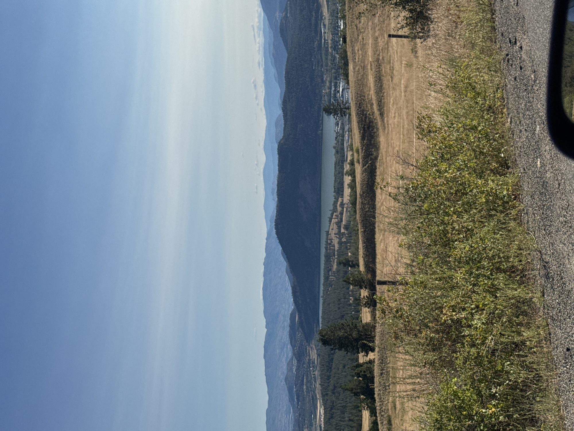 The image captures a panoramic view of a valley landscape near Lake Country, Canada, under a clear blue sky with sparse, wispy clouds. The foreground is dominated by a rough gravel road surface, partially visible in the bottom right corner, and dense, green foliage with dry, brown grasses and brush. A wooden fence post stands prominently in the middle ground.
Beyond the foreground, rolling hills covered in dry, golden grasses stretch out. A dense forest of dark green coniferous trees covers the slopes of the hills. In the mid-distance, a serene lake is visible, reflecting the sky and surrounding landscape. The far distance is characterized by layers of progressively faded blue mountains, creating a sense of depth and vastness. A small settlement or town is nestled near the lake, indicated by scattered buildings and structures. The overall scene depicts a natural, semi-arid environment with signs of human habitation.
