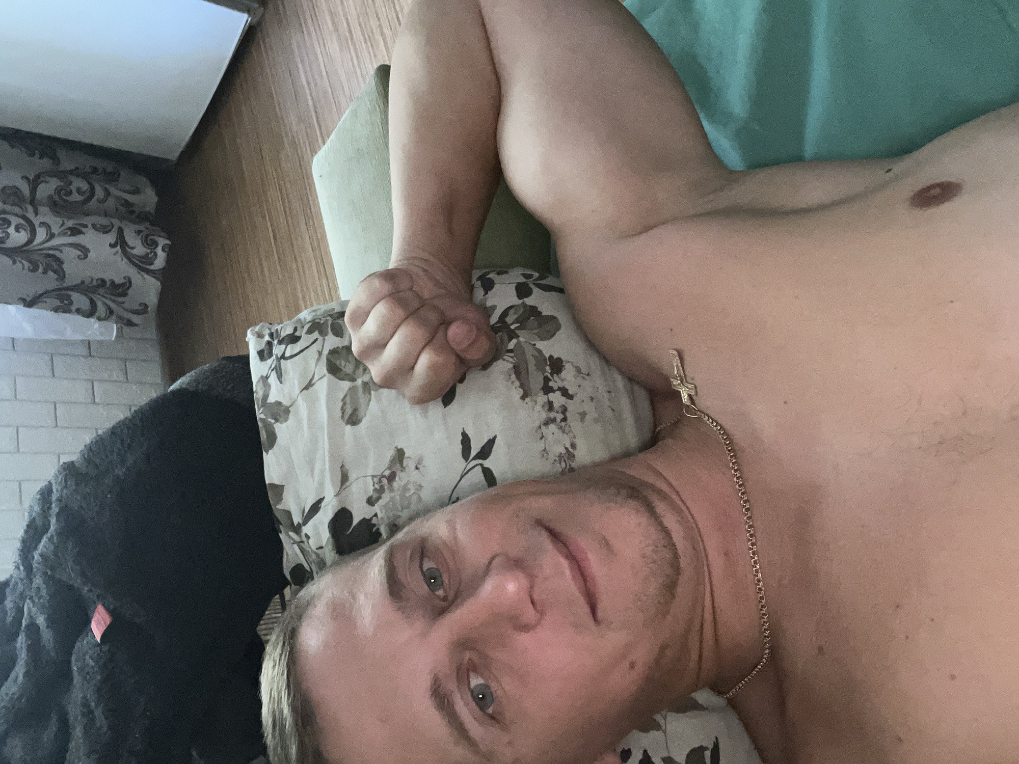 A fair-skinned man with light brown hair and blue eyes is lying on his back, with his head resting on a floral-patterned pillow. He is looking directly at the camera and smiling slightly. His left arm is bent at the elbow, with his fist clenched and resting on the pillow. His right arm is extended along his side. He is shirtless and wearing a thin gold chain with a cross pendant. The background shows a wall with light-colored horizontal bricks and a patterned curtain on the right. The floor is a wooden laminate. The scene appears to be indoors, in a residential setting. Based on the provided context, the location is the city of Minsk, Belarus.