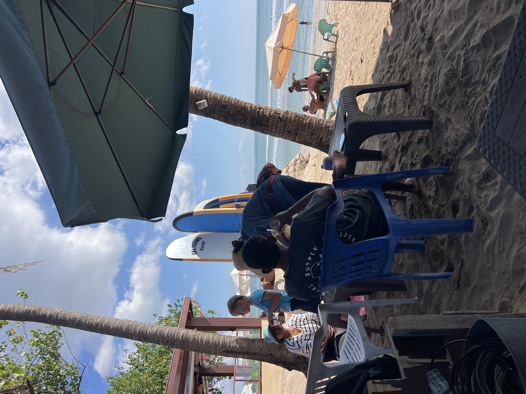 In Kuta, Indonesia, a beach scene unfolds under a bright, partly cloudy sky. Several people are present, some seated at tables on the sand and others gathered near the water. In the foreground, a person in a black t-shirt with "SUPPLY" printed on it sits in a blue chair at a dark plastic table. To their left, a person in a striped shirt and a light blue hat sits in a white chair. A young girl with braided hair, wearing a blue shirt, stands near them, holding a drink.
Behind the seated individuals, two surfboards stand upright on the sand, one white with "BALIGUNS SURF" written on it, and a blue one next to it. A palm tree stands prominently, its trunk textured and reaching towards the sky. Further back, a large green umbrella shades part of the seating area.
On the beach, closer to the water, a beige umbrella provides shade for a few people seated on chairs. Two men appear to be conversing. A few more individuals are visible near the ocean, with gentle waves rolling onto the shore. The overall environment is a relaxed beach setting, likely a beachfront cafe or relaxation spot.