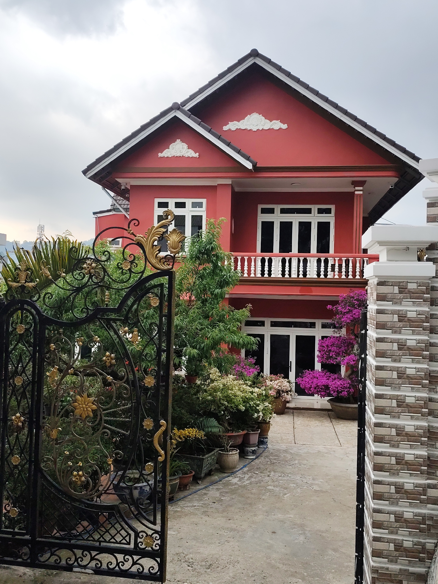 The image presents a vibrant residential property in Cam Ly - Đà Lạt, Vietnam, centered around a striking two-story house with a prominent red exterior. The house features a dark-tiled, pitched roof, adorned with white trim and decorative white plasterwork on the gables. White-framed windows and doors, some with dark interiors, are visible on both floors. A white balustrade adorns the second-story balcony.
In the foreground, a partially open, ornate wrought-iron gate with intricate black scrollwork and golden floral accents provides a grand entrance. Beyond the gate, a paved pathway leads toward the house, bordered by a lush and well-maintained garden. Various green trees, shrubs, and numerous potted plants, including vibrant fuchsia bougainvillea and other flowering plants in white and pink hues, line the path and surround the house's base.
On the right, a decorative wall or pillar constructed from multi-toned gray and brown tiles with a textured finish partially frames the scene. The sky is overcast, suggesting a cloudy daytime. No people or visible text are present in the image. The scene conveys a sense of well-kept beauty and peaceful residential living.