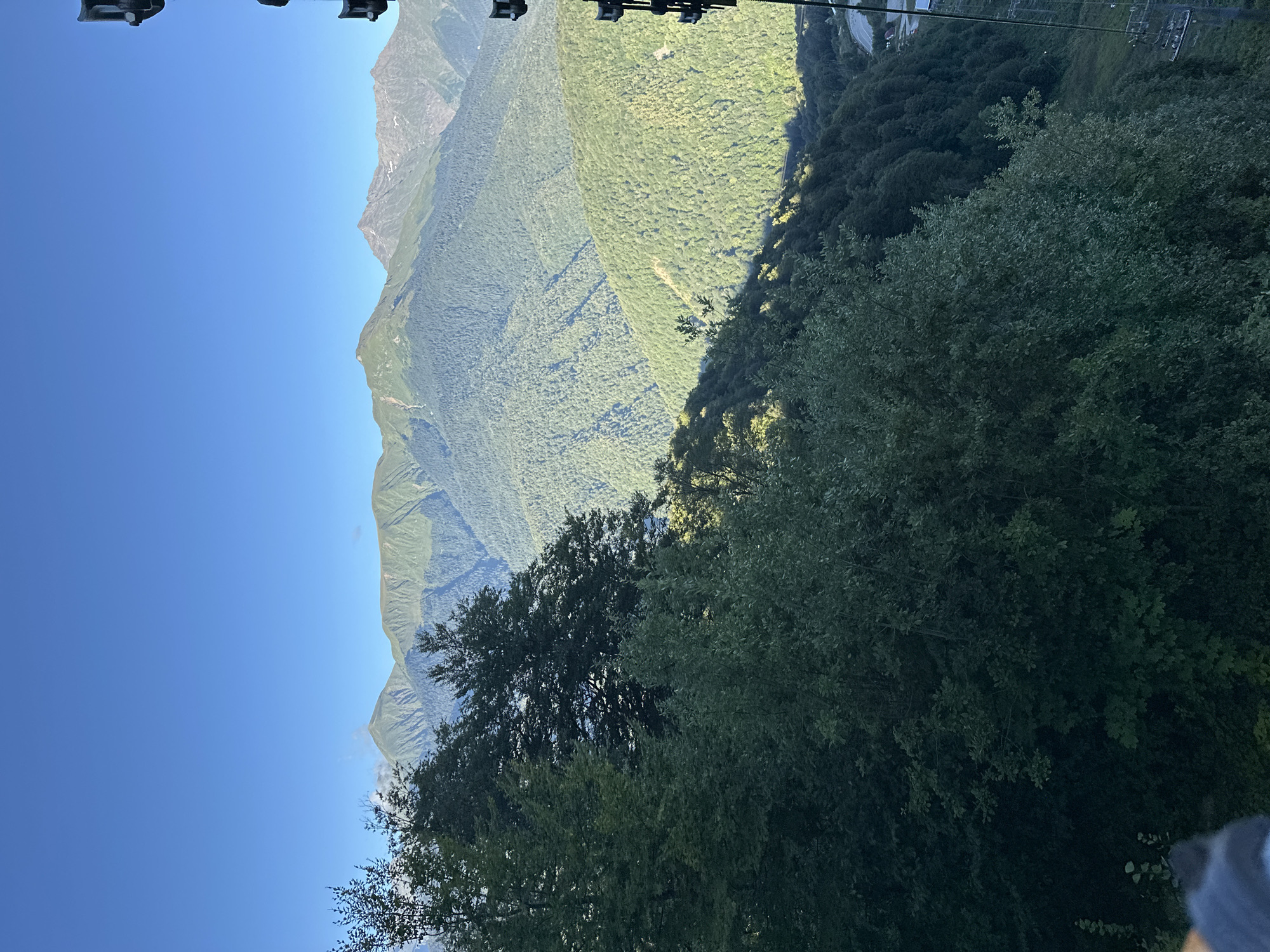 This is a vertical, outdoor, eye-level photograph taken from a high vantage point, likely a mountain or ski lift, overlooking a vast mountain range and dense forest in Estosadok, Russia. The sky is a clear, vibrant blue, with no clouds visible. In the foreground, lush green trees dominate the left side of the frame, with some branches extending into the lower portion of the image. Behind the trees, a series of undulating green mountain slopes recede into the distance, covered in thick, dark green forest. The sunlight casts shadows and highlights on the mountains, creating a sense of depth and texture. In the far distance, the peaks of larger mountains are visible, some with patches of snow. On the far right edge of the image, part of a black ski lift apparatus is visible, with a few empty chairs hanging from it, suggesting a resort area. The overall scene conveys a sense of natural beauty and tranquility, typical of mountainous landscapes in the summer season.