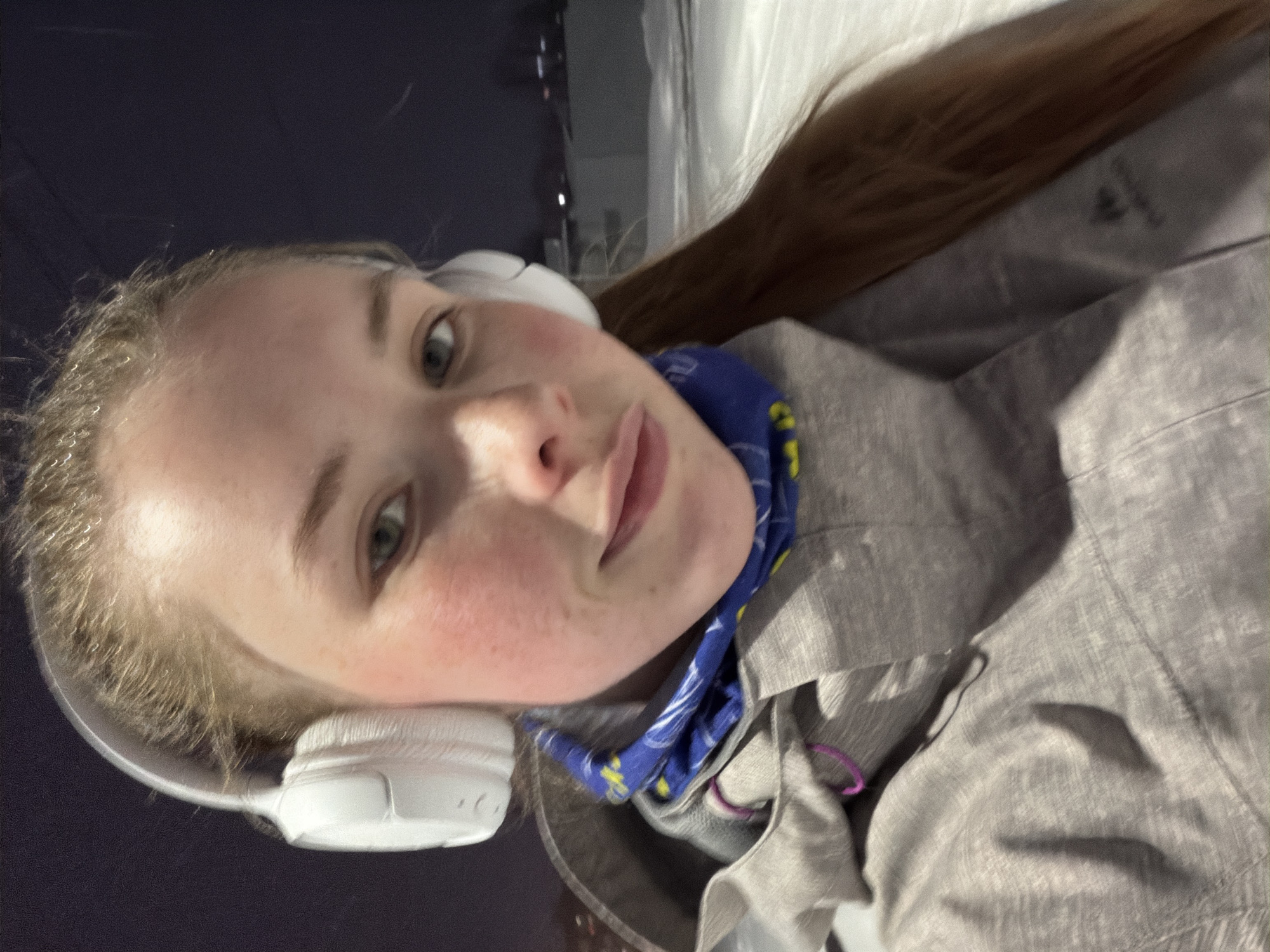 A young woman wearing white headphones and a blue patterned neck gaiter poses for a selfie. She is wearing a gray jacket. The background is dark, possibly nighttime, with a suggestion of snow or a light-colored structure. There is nothing present in the image to confirm the location as the city of Berezniki, Russia.