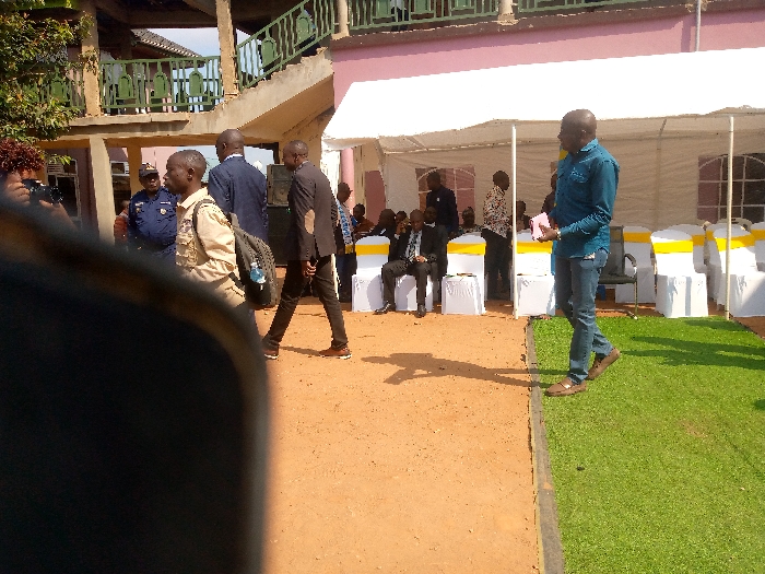 The image shows a gathering of people outside a building in Beni, Democratic Republic of Congo. The location, based on the provided coordinates, is a residential area with a mix of buildings and open spaces. The scene is set up for a formal event, with a white tent and rows of white chairs with yellow sashes. Several people are seated in the chairs, while others are standing around, some in conversation. A man in a blue shirt and jeans is holding a piece of paper and appears to be speaking to someone off-camera. Another man is standing in the background, taking a photo with a camera. The building in the background has a pink wall and a green roof, with stairs leading up to the second floor. The ground is a mix of dirt and grass, with a clear line dividing the two surfaces. The overall atmosphere is one of anticipation and excitement for the event.
