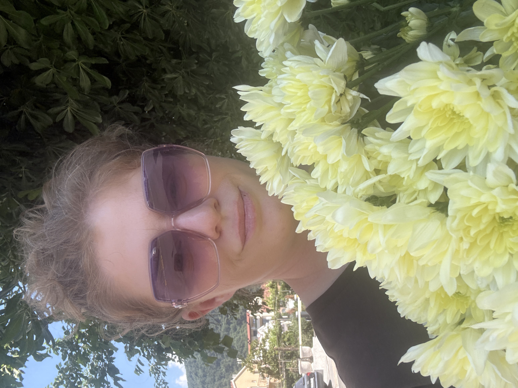 A woman is holding a bouquet of light yellow chrysanthemums. She is wearing sunglasses with a purple tint. Her hair is light-colored and curly. The background shows buildings and a hillside, suggesting an urban environment. Based on the architecture and landscape, this location is likely in Budva, Montenegro.
