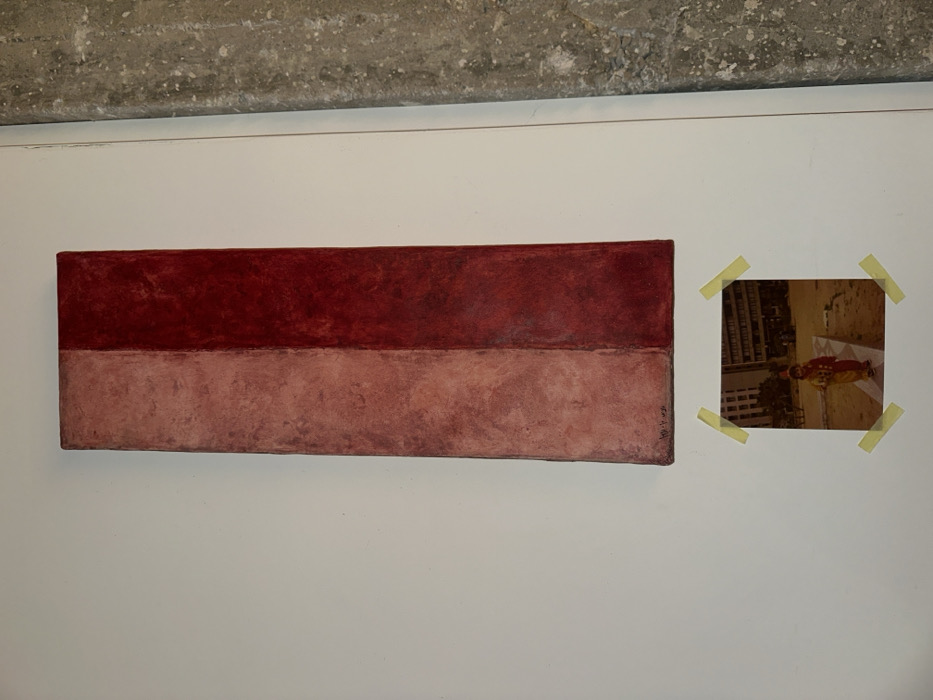 On a white wall with a concrete section above, a rectangular artwork is displayed. The piece features two horizontal color blocks: a darker maroon above and a lighter, almost pinkish-brown below. To the right, a square photograph is affixed to the wall with yellow tape at each corner. The photograph appears to show an individual in a colorful outfit, possibly lying on the ground, with a cityscape in the background.