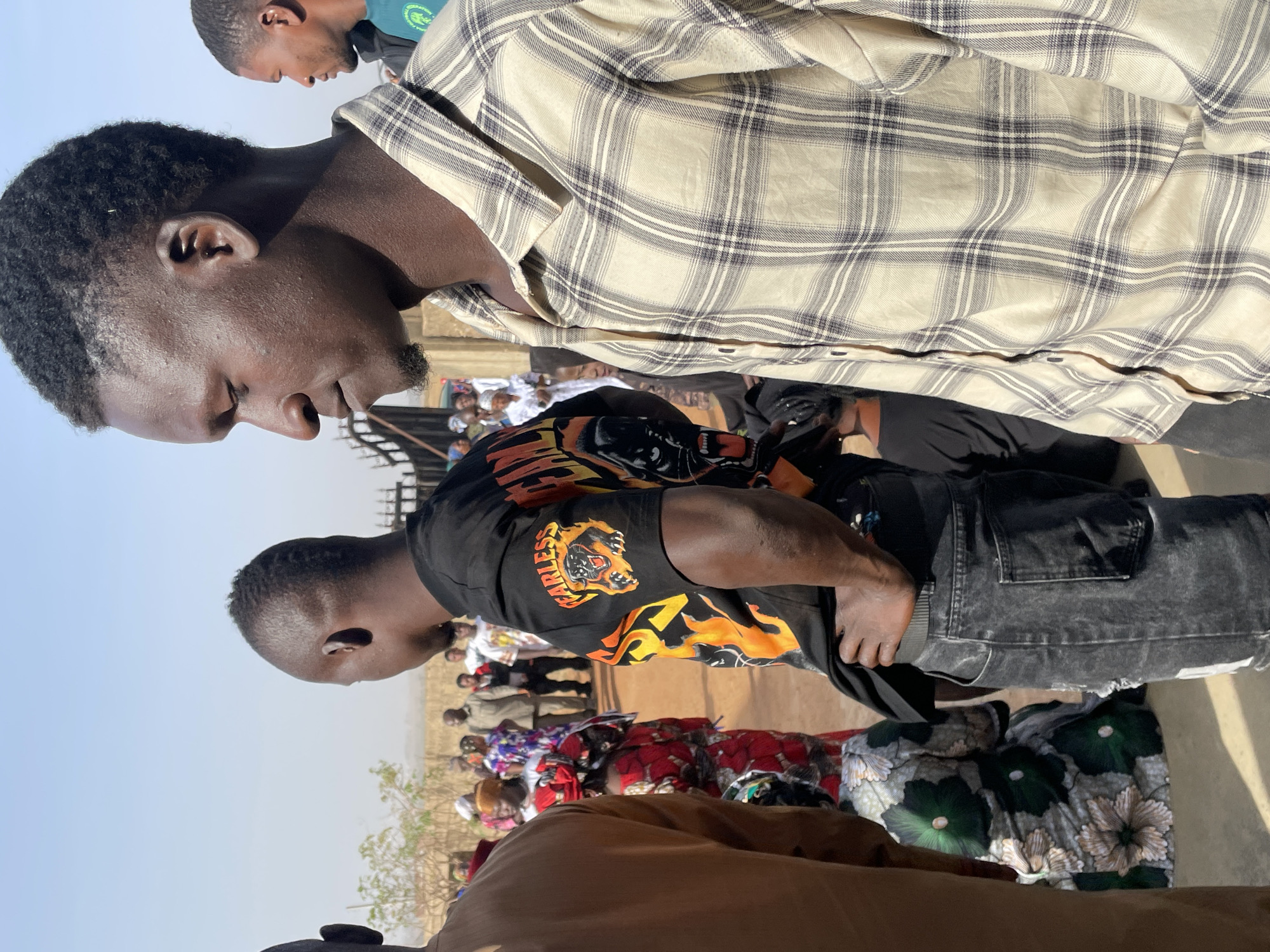 This outdoor image, taken in Du, Nigeria, captures a gathering of people under a clear, bright sky. The scene is dominated by several Black men in the foreground and midground, with more individuals, including women in colorful traditional attire, visible further back.
In the immediate foreground, a man with short, dark, curly hair and a subtle goatee is seen in profile, looking downwards with a focused expression. He wears a light beige plaid shirt with dark grey lines. Behind him, another man with short, dark hair is also seen in profile, his face partially obscured, wearing a dark green shirt.
The midground features a man with short hair, viewed from the back and side, wearing a black T-shirt adorned with a prominent orange and yellow graphic. The graphic depicts a roaring animal head, possibly a tiger or lion, with the word "FEARLESS" written in orange above it. He also wears dark, possibly distressed, jeans. His posture suggests he might be looking at something on the ground or in front of him, with his left hand near his waist.
In the background, a crowd of people is visible, some standing, some possibly seated on the ground, with women wearing vibrant, patterned fabrics. The ground appears to be unpaved, suggesting an open community area or market. The strong sunlight and lack of clouds indicate a clear day, likely midday or early afternoon. No specific event is clearly identifiable, but it appears to be a social gathering or a public market setting.