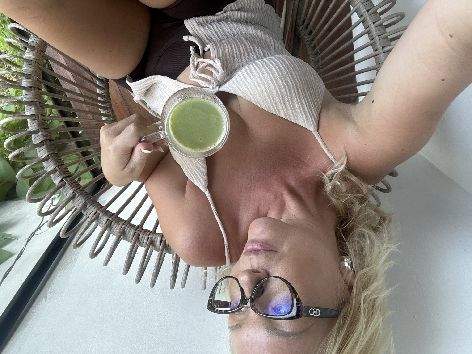 A blonde woman with fair skin and a light tan sits in a wicker chair holding a clear mug filled with a green beverage. She is wearing a light-colored, textured camisole with thin straps and dark brown shorts. She also wears large, cat-eye glasses with a tortoiseshell pattern and silver hoop earrings. The background appears to be an outdoor setting, possibly a balcony or patio, with a white wall and greenery visible behind the chair. The lighting suggests it is daytime.