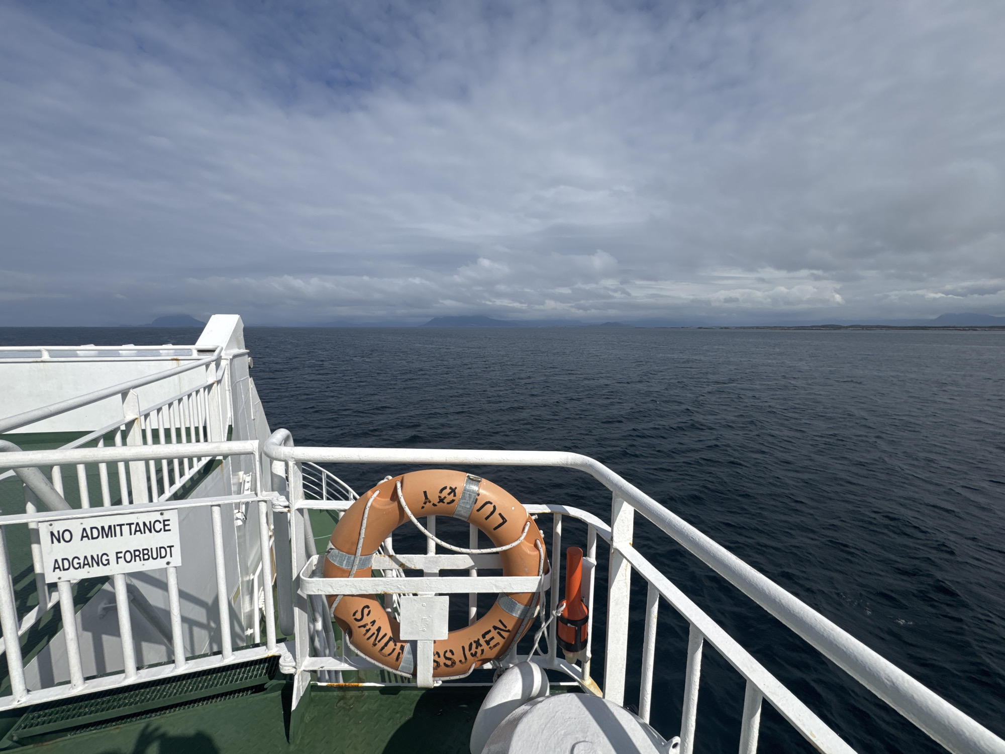 The image captures a view from a ferry deck, looking out at the ocean under a cloudy sky. A white railing runs along the left and right sides of the frame, separating the deck from the water. A life preserver, bright orange with white lettering, is mounted on the railing on the right side. A white sign with black text reading "NO ADMITTANCE ADGANG FORBUDT" is attached to the railing on the left. The water is a dark blue, with small ripples indicating a gentle swell. In the distance, a faint line of land with hills or mountains is visible on the horizon. The ferry appears to be in motion, suggesting it is traveling. This scene is likely taking place in the waters surrounding Sleneset, Norway, a coastal area known for its ferry services and maritime landscape.