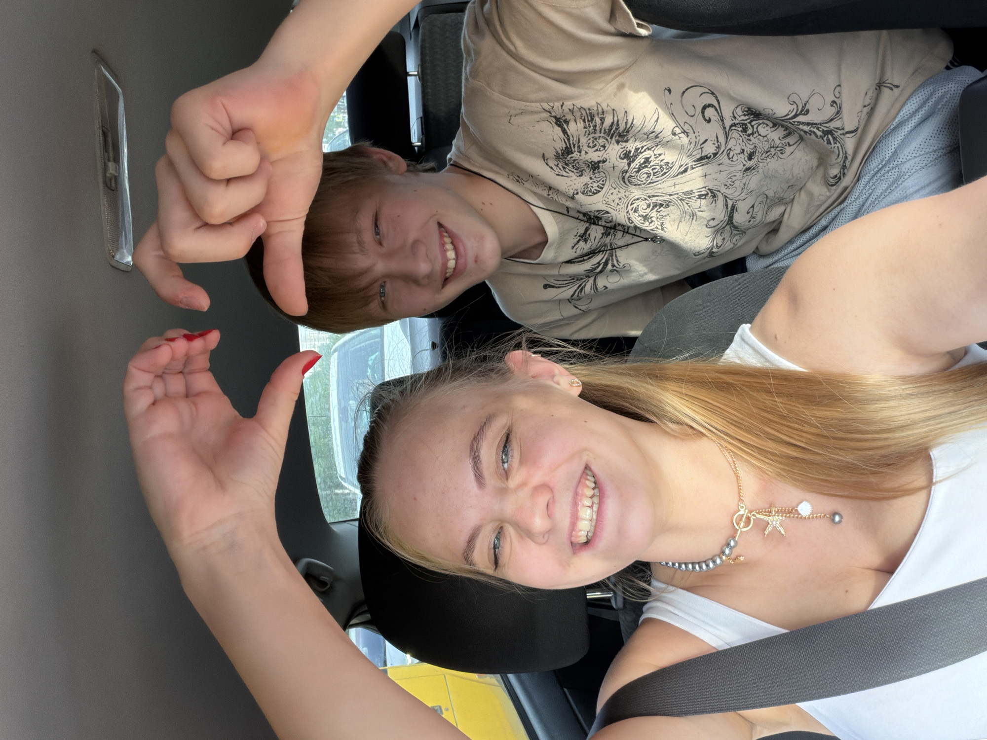 Two people are in a vehicle. A fair-skinned young woman with blonde hair, wearing a white top, sits in the driver's seat and smiles at the camera. She is wearing a seatbelt and a necklace with a star pendant. Her nails are painted red. She raises her right hand, index finger extended. A fair-skinned young man with brown hair, wearing a patterned beige shirt, sits behind her. He smiles and makes a gesture with his hand. The interior of the vehicle is visible. Through the window, a blurred view of the outside includes a yellow vehicle and other cars. The location is Izhevsk, Russia.