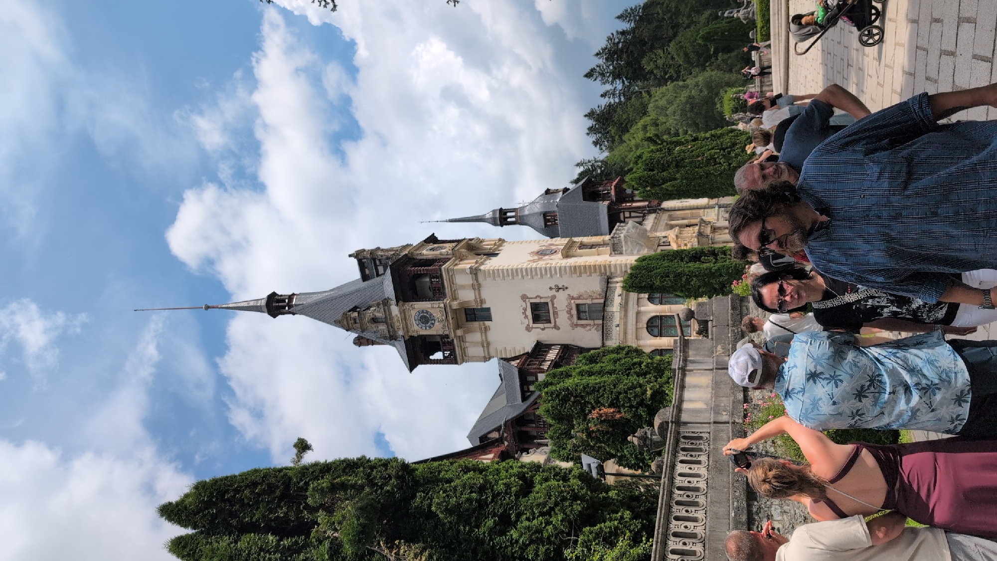 This image shows a view of Peles Castle in Sinaia, Romania. The castle is a magnificent structure with a tall central tower topped by a spire, and several smaller turrets. It is surrounded by lush green trees and a partly cloudy blue sky. In the foreground, a group of tourists are walking on a paved path. A woman in a burgundy dress is taking a picture with her phone, while a man in a blue patterned shirt is looking forward. Another man in a blue striped shirt is in the right foreground, looking to the side. A baby in a stroller is also visible in the background on the right. The scene suggests a warm, sunny day at a popular tourist destination.