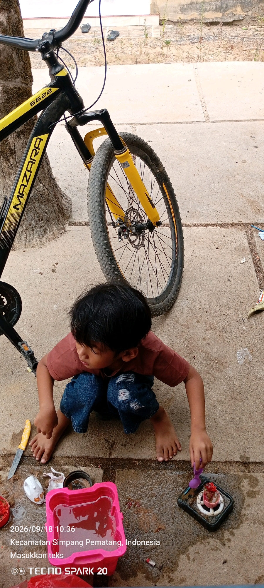 A young child with dark hair, wearing a brown t-shirt and distressed blue jeans, is crouched on a paved surface next to a bicycle. The child's back is partially to the camera, and they are looking down at their hands, which are near a pink container filled with reddish liquid and a small black tray with a purple spoon. The bicycle, predominantly black with yellow accents, has its front wheel in the foreground, obscuring part of the child. The background consists of a tree trunk, a light-colored wall, and a sandy area with scattered debris. The scene is dated "2026/09/18 10:36" and identified as being in "Kecamatan Simpang Pematang, Indonesia."