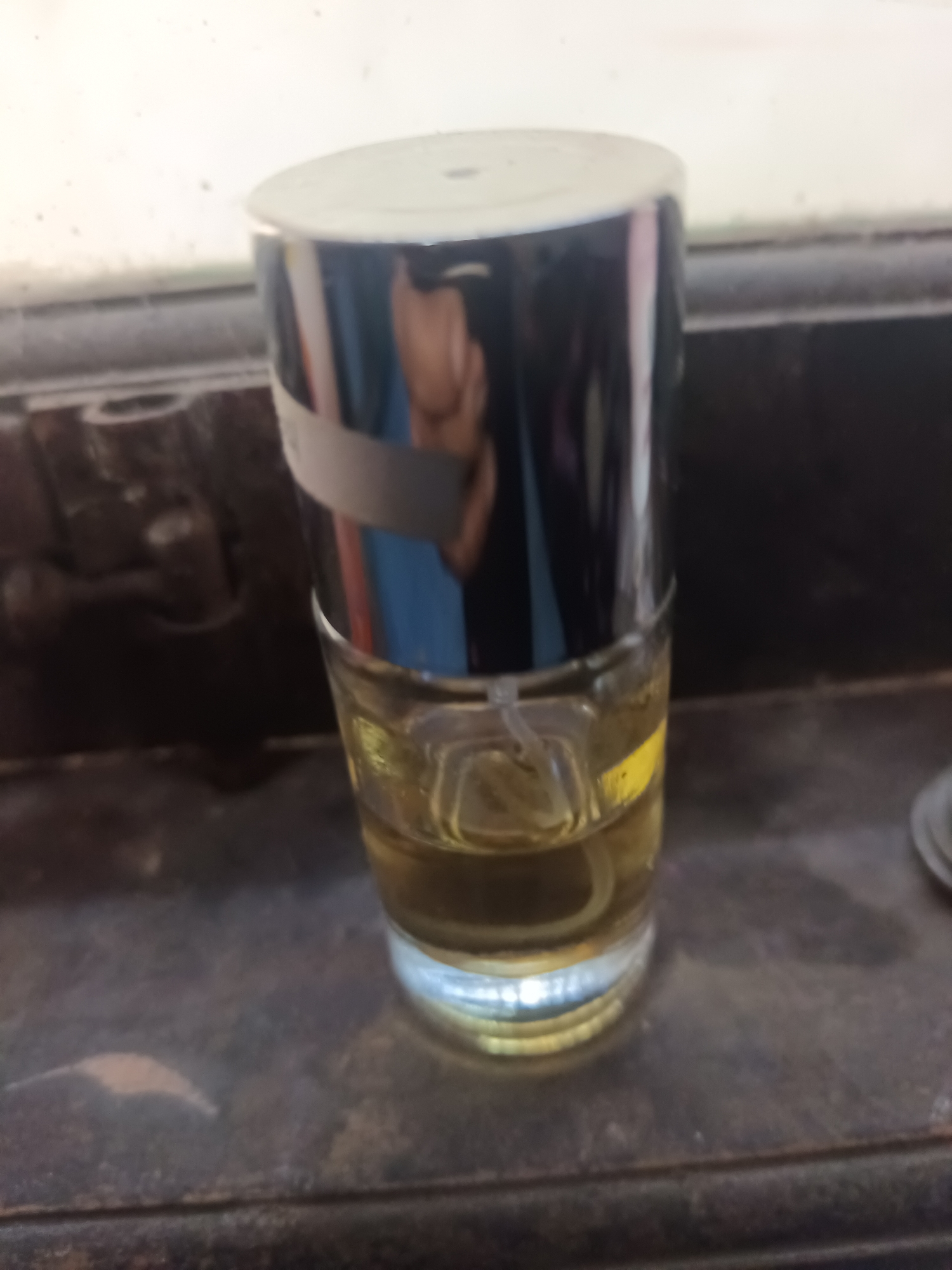 A partially filled perfume bottle with a silver cap and a clear glass base is positioned on a dark, textured surface. The liquid inside the bottle is a light yellow color. The background is out of focus and appears to be a wall with some lighter colored sections. The scene is indoors, and the lighting suggests artificial light. No people or specific urban landmarks are visible to indicate a location in Terbanggi Marga, Indonesia.