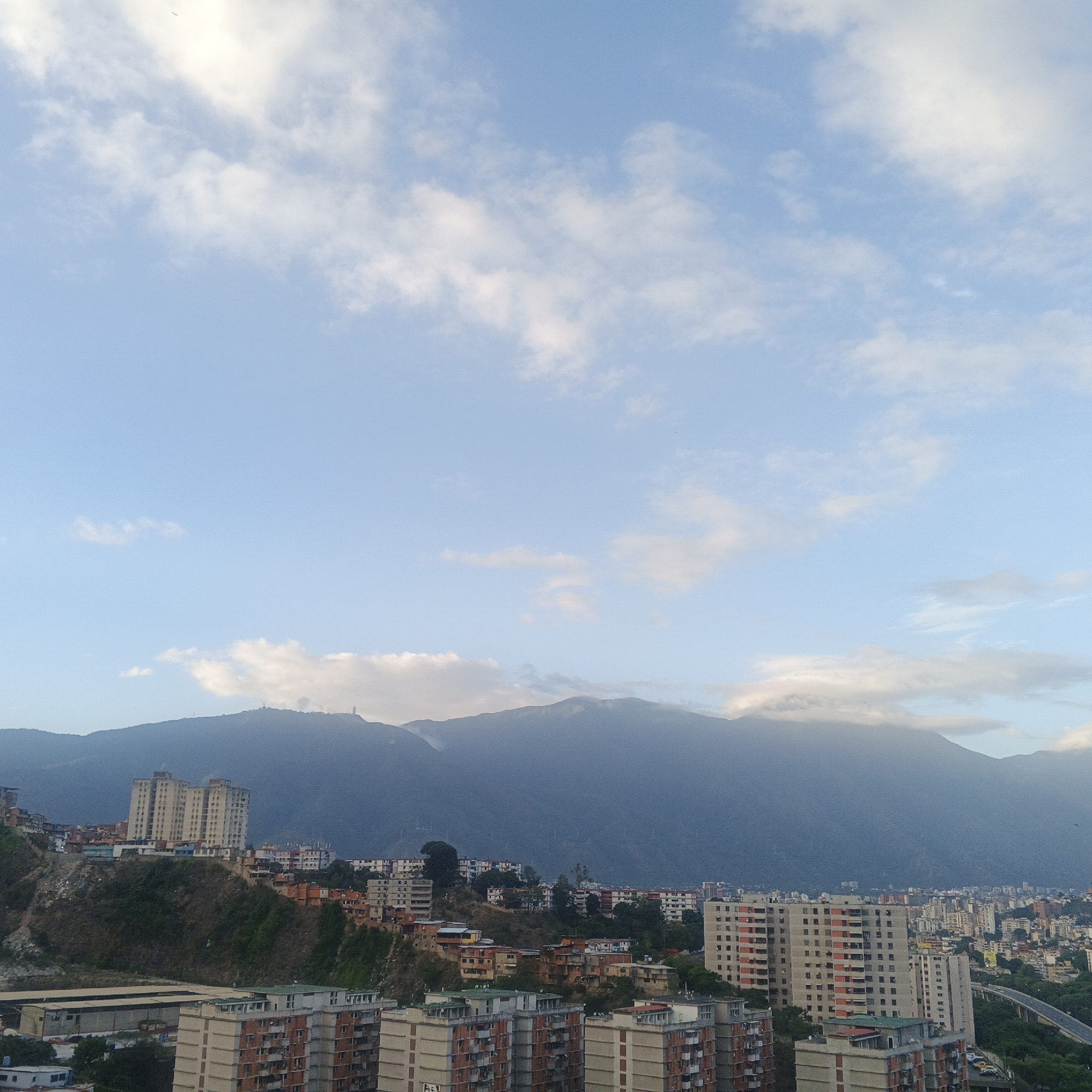 This image captures a panoramic view of a densely populated urban area nestled against majestic mountains, characteristic of Caracas, Venezuela. The scene is dominated by a vast, light blue sky scattered with white and light gray stratocumulus clouds, suggesting either early morning or late afternoon.
In the background, a formidable range of mountains rises, partially shrouded in a soft, misty haze or low-lying clouds that cling to their peaks. On the ridge of the left-most mountain, a prominent slender structure, possibly a communication tower or monument, is visible, with other smaller towers discernible along the crest.
The city itself stretches across the midground and foreground, built upon undulating hillsides. A mix of multi-story apartment buildings, varying in architectural style and age, dot the landscape. Many structures have a utilitarian, concrete appearance. On the steeper slopes, numerous smaller, colorful houses are clustered together, indicative of informal settlements or dense neighborhoods. Patches of green vegetation are visible on the hills and between the buildings. Towards the lower right, an elevated road or bridge curves through the urban sprawl, indicating infrastructure connecting different parts of the city.
The overall impression is one of a vast urban landscape coexisting with impressive natural scenery under a bright, yet softly lit sky. No human activity or visible text is explicitly discernible in the frame.