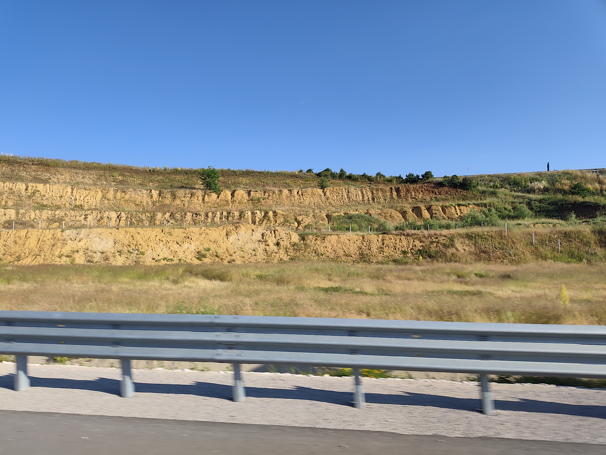 The image depicts a roadside view in Brestnitsa, Bulgaria, featuring a barren, tiered hillside adjacent to a road. The hillside is characterized by distinct horizontal layers of exposed earth, predominantly yellow and orange in color, with patches of dry grass and sparse vegetation. Several small trees are scattered across the top of the hill. In the foreground, a gray metal guardrail runs parallel to the road, supported by concrete posts. The road surface is visible below the guardrail. The sky above is a clear, bright blue with a faint contrail visible. The scene appears to be captured on a sunny day, possibly from a moving vehicle, given the slight motion blur.