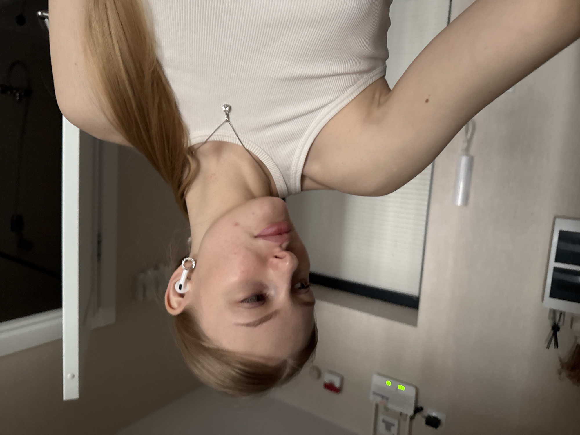 The image shows a young woman with long blonde hair, wearing a white tank top and an earbud with a sparkly hoop. She appears to be inside a room with beige walls. A window with blinds is visible in the background, along with a bathroom vanity and other unidentified wall fixtures. There is no information available in the image to determine the location as the city of Perm, Russia. The image provides no information about actions, interactions, or specific subjects beyond the woman herself.