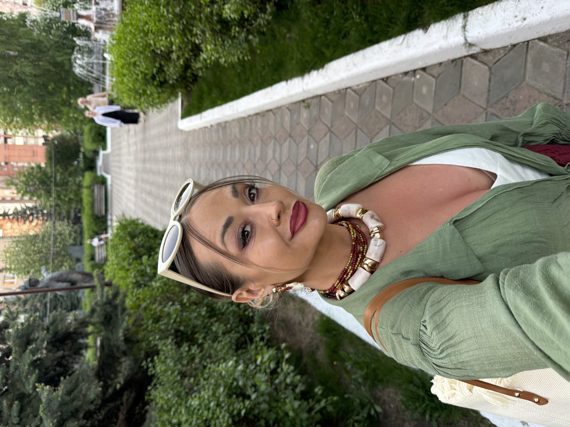 A female subject is depicted in a close-up selfie, positioned slightly off-center to the right, in a public park in Krasnoyarsk, Russia. She has medium-brown hair, dark eye makeup, and dark red lipstick. White-framed sunglasses are pushed up onto her head. She wears gold-toned dangle earrings and a layered necklace featuring a string of small red and gold beads topped by a strand of large, irregular white stones with gold accents. Her attire consists of an open olive-green, textured long-sleeved shirt over a white or cream top, revealing cleavage. A light-colored woven bag with a brown leather strap is visible on her left shoulder.
The background shows a paved pedestrian pathway constructed of geometric grey and light-colored interlocking pavers, bordered by a white concrete curb. Lush green bushes line both sides of the pathway. On the left, coniferous trees and a dark-colored statue of a standing male figure are visible. Further down the path on the left, a person is seated on a bench. On the right side, deciduous trees and a fountain actively spraying water are present in the middle distance. Two individuals are walking away from the camera near the fountain: one in a dark dress and white overshirt, and another in light-colored clothing. Multi-story buildings with reddish facades are discernible in the distant background, beyond the park's trees. The scene is illuminated by soft, diffused light, suggesting late afternoon or early evening.