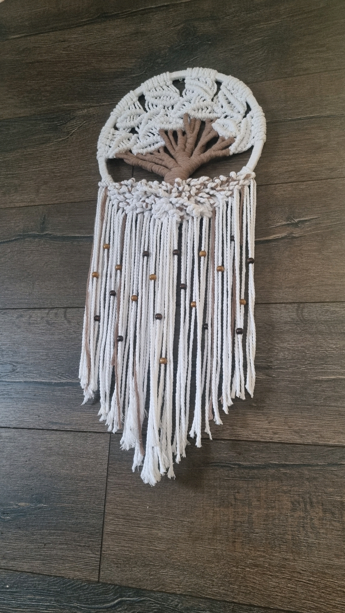 The image shows a macrame wall hanging with a tree of life design against a dark wooden background. The wall hanging is predominantly white with brown accents. The tree of life is depicted within a circular frame, with the trunk and branches in brown and the leaves in white macrame. Below the tree, there are multiple hanging cords, some of which are adorned with small wooden beads. The wooden background appears to be floorboards or a wall paneling. The scene is static, with no people or animals present. The lighting suggests an indoor environment. There is no information available to determine the location within the city of Kyiv, Ukraine.