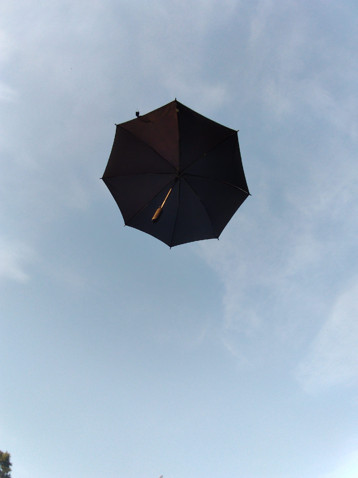 The image shows a black umbrella floating in a pale blue sky with wispy clouds. The umbrella is open and viewed from below, with its handle and a portion of its shaft visible. The fabric of the umbrella appears to be dark, possibly black or a very dark shade of another color. The umbrella is centrally located in the frame, suspended against the sky. The sky is bright, suggesting it is daytime. There are no people visible in the image. The scene appears to be outdoors, with the sky as the primary background. The location context provided is Chaikovsky, Russia, but there are no specific visual cues in the image to confirm or deny this. The overall impression is one of an unusual event, with an object that normally protects from precipitation unexpectedly airborne. There is no visible text in the image.
