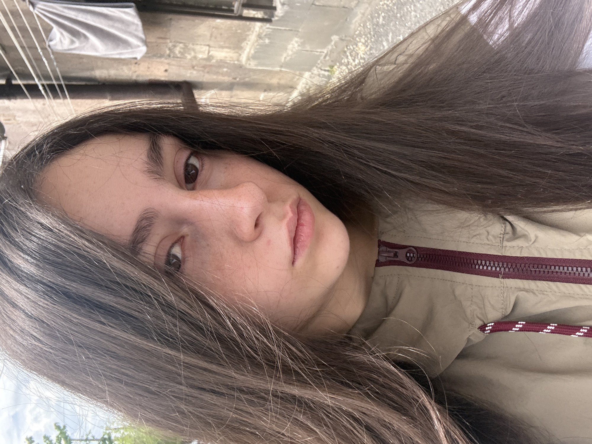 A close-up, angled portrait of a young woman with long, dark brown hair. She has fair skin, brown eyes, and freckles on her face. She is wearing a light brown jacket with a maroon zipper and drawstrings visible near her neck. The background shows an urban scene, with concrete structures and a clothesline with a piece of laundry hanging above, suggesting Yerevan, Armenia. The ground appears to be paved with stone.