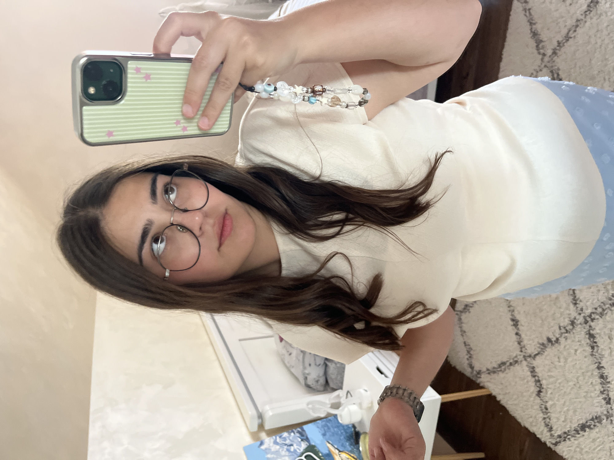 A young woman with long brown hair and glasses takes a selfie in a bedroom. She is wearing a cream-colored top and a light blue skirt. She is holding up a smartphone with a pastel green case decorated with white stripes and pink stars. A beaded phone charm hangs from the phone. The background shows a white bed, a dresser with items on top, and a patterned rug on the floor. The room appears to be a domestic setting. The image was captured in Koroviia, Ukraine.