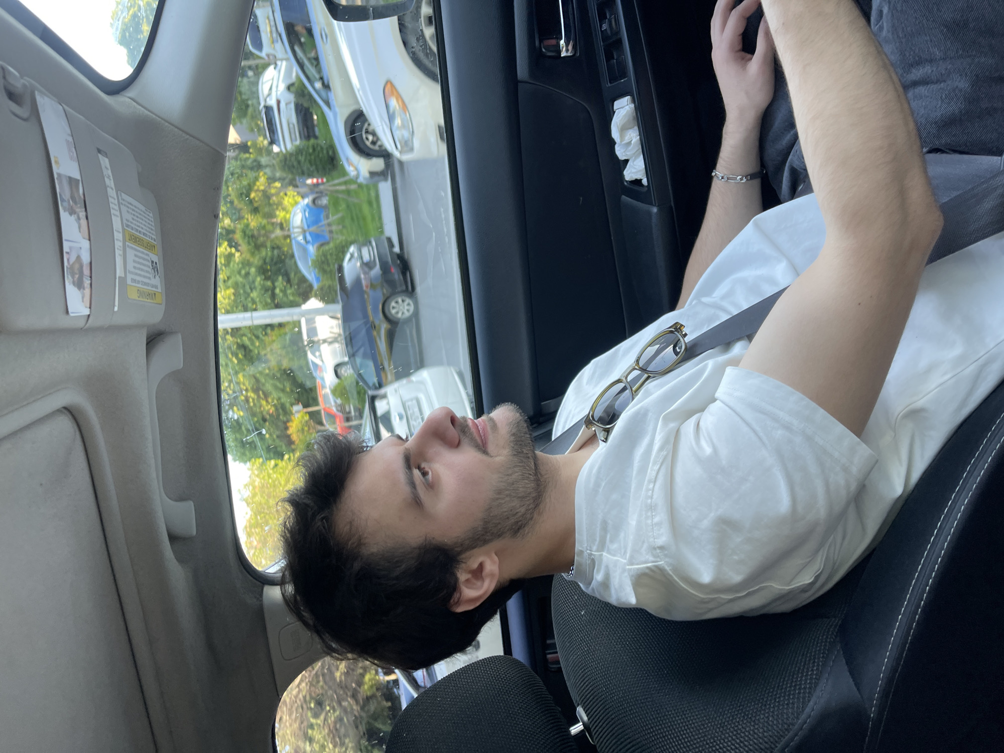 The image depicts a man seated in the driver's seat of a car. He has dark hair and a beard, and he is wearing a white shirt. A seatbelt is buckled across his chest. Sunglasses are placed on top of the shirt. The car appears to be stationary, as other vehicles are visible through the window. Based on the visible vegetation and architectural style, the location might be the city of Tbilisi, Georgia. The interior of the car features a black dashboard and a grey interior.