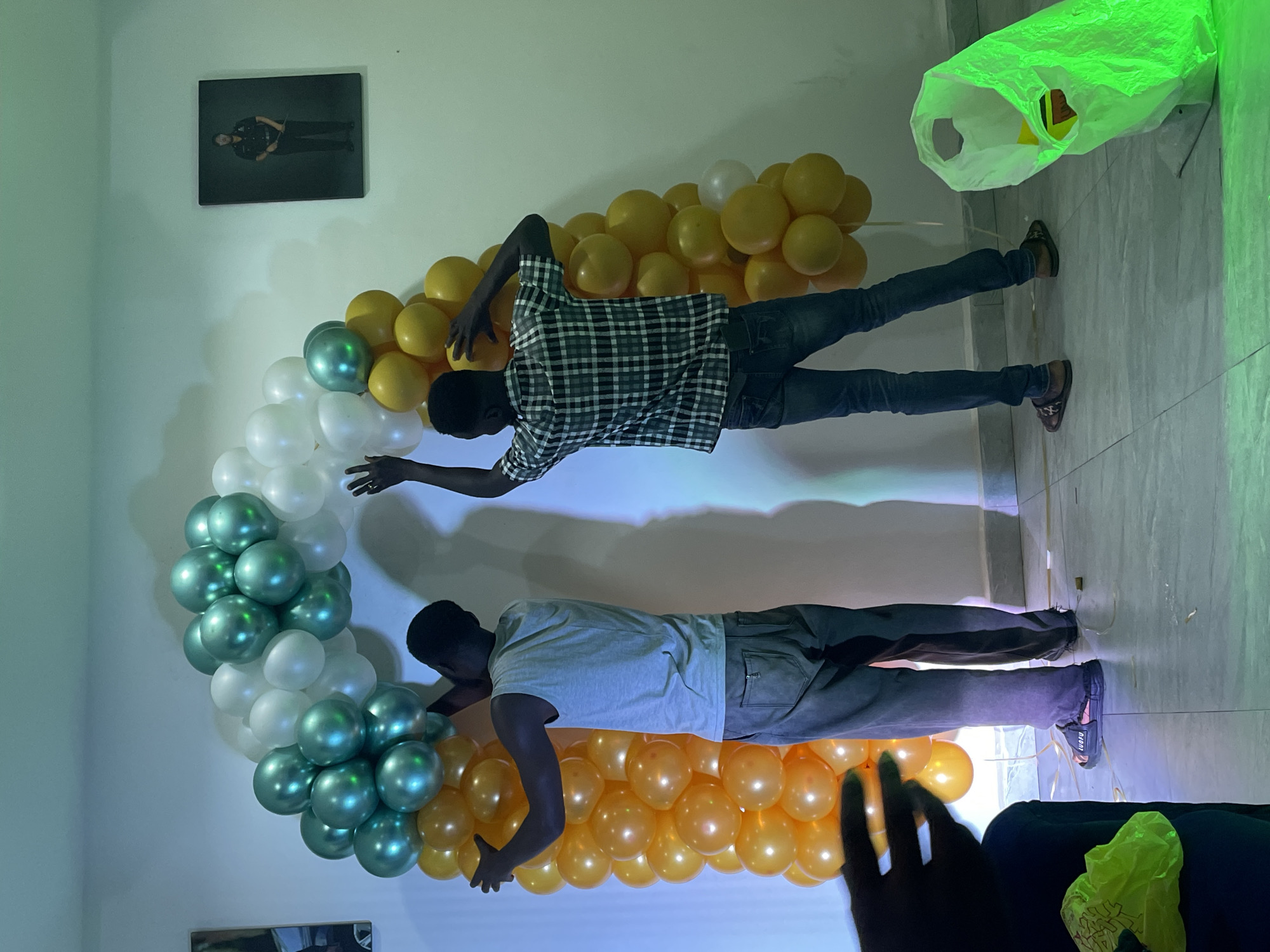 Two individuals are in the process of constructing a balloon arch in front of a wall. The arch consists of balloons in green, white, and orange hues, forming a semicircular structure. One person, wearing a light-colored tank top and grey pants, is positioned on the left side of the arch, arranging balloons. The other individual, dressed in a checkered shirt and blue jeans, stands on the right, also contributing to the balloon arrangement. The background features a white wall with a framed picture of a person in uniform hanging on it. The floor is tiled, and there is a bright green plastic bag in the foreground on the right side of the image. The lighting in the room appears to be artificial, casting shadows and creating a festive atmosphere.