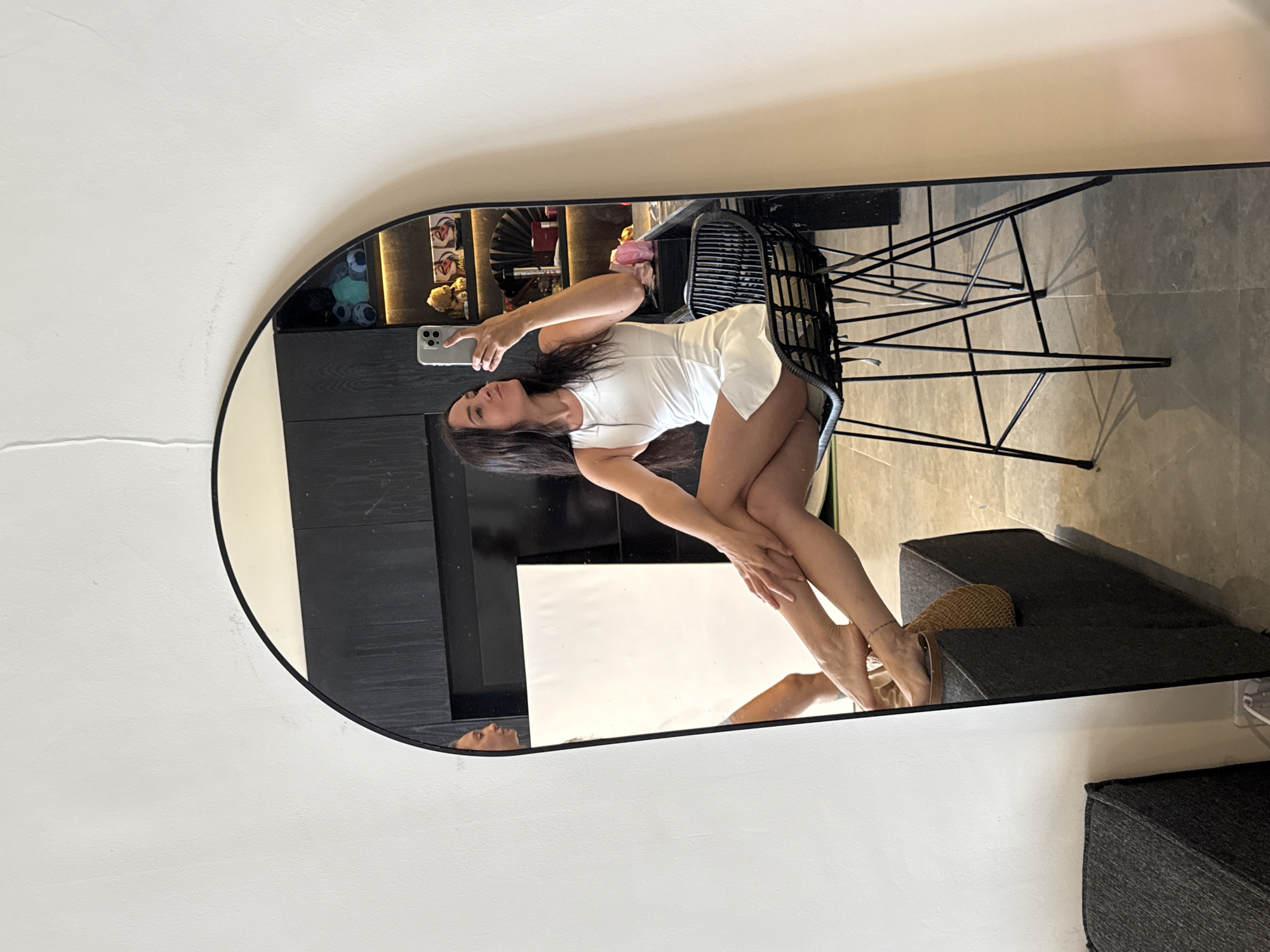 A woman is taking a selfie in a mirror in what appears to be a modern, well-lit room. She is wearing a white sleeveless dress and is seated on a black metal chair with a woven seat. Her legs are crossed, and she is holding her phone up to take the picture. Her expression is playful, with her lips pursed.
The mirror is large and arched, reflecting a significant portion of the room. Behind her, shelves are visible with various decorative items, including stuffed animals and pictures. A black wooden cabinet is also present. In the reflection on the left side of the mirror, another person's face is visible, looking away from the camera.
The floor appears to be tiled with a light gray pattern. To the left of the mirror, a dark gray upholstered couch is partially visible. The overall impression is one of a stylish and comfortable living space. The prompt does not provide specific information about the location being in North Kuta, Indonesia, so this cannot be confirmed from the image alone.