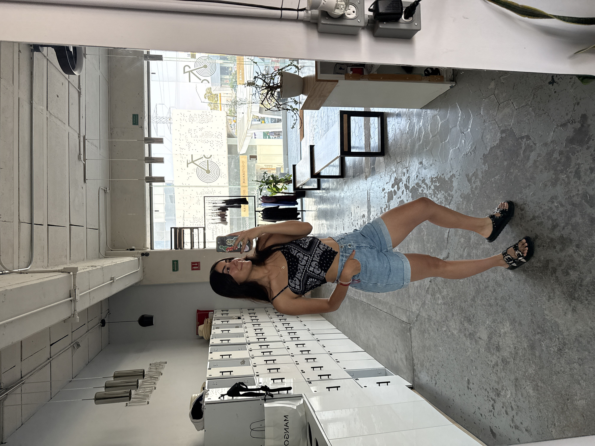 A young woman is seen taking a selfie in what appears to be a reflection in a large mirror inside the 'BAINA STUDIO' in the city of Puebla, Mexico. The background includes lockers, a bench, and a cityscape visible through a large window. The floor is grey. The woman is wearing a paisley crop top and denim shorts.