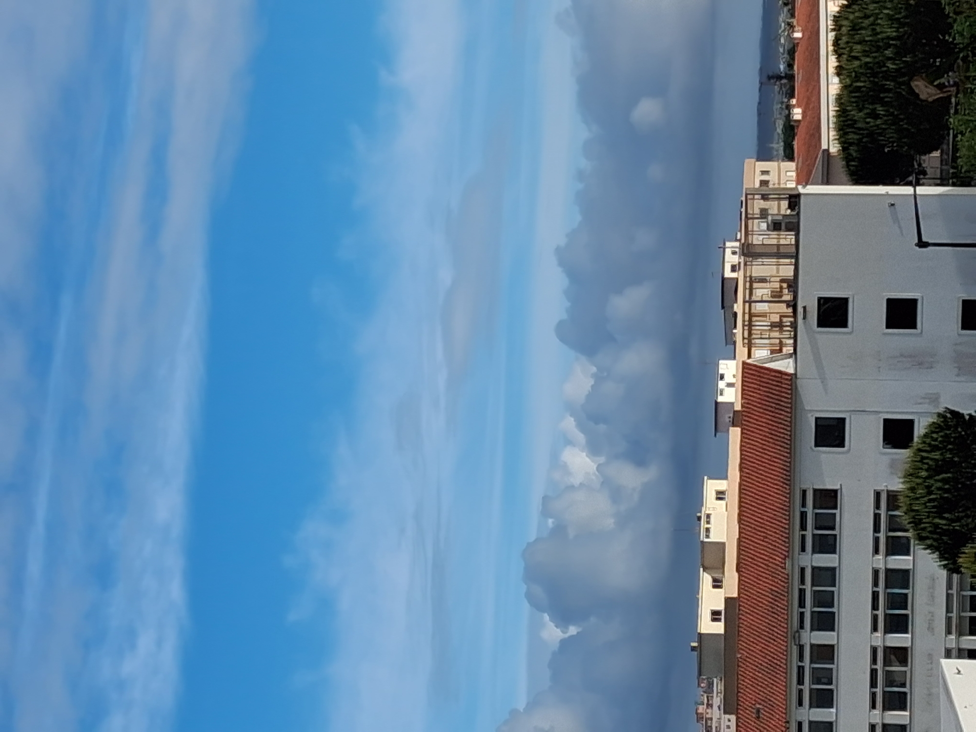 The image presents a vertical view of a scene in Figueira da Foz, Portugal. On the right, buildings are visible, with light-colored facades, some featuring red-tiled roofs and windows. The urban environment includes greenery in the form of trees or bushes. The left side of the image is dominated by the sky, featuring a mix of blue sky and clouds. The clouds vary in density, ranging from thin, wispy formations to denser, darker cumuliform clouds.