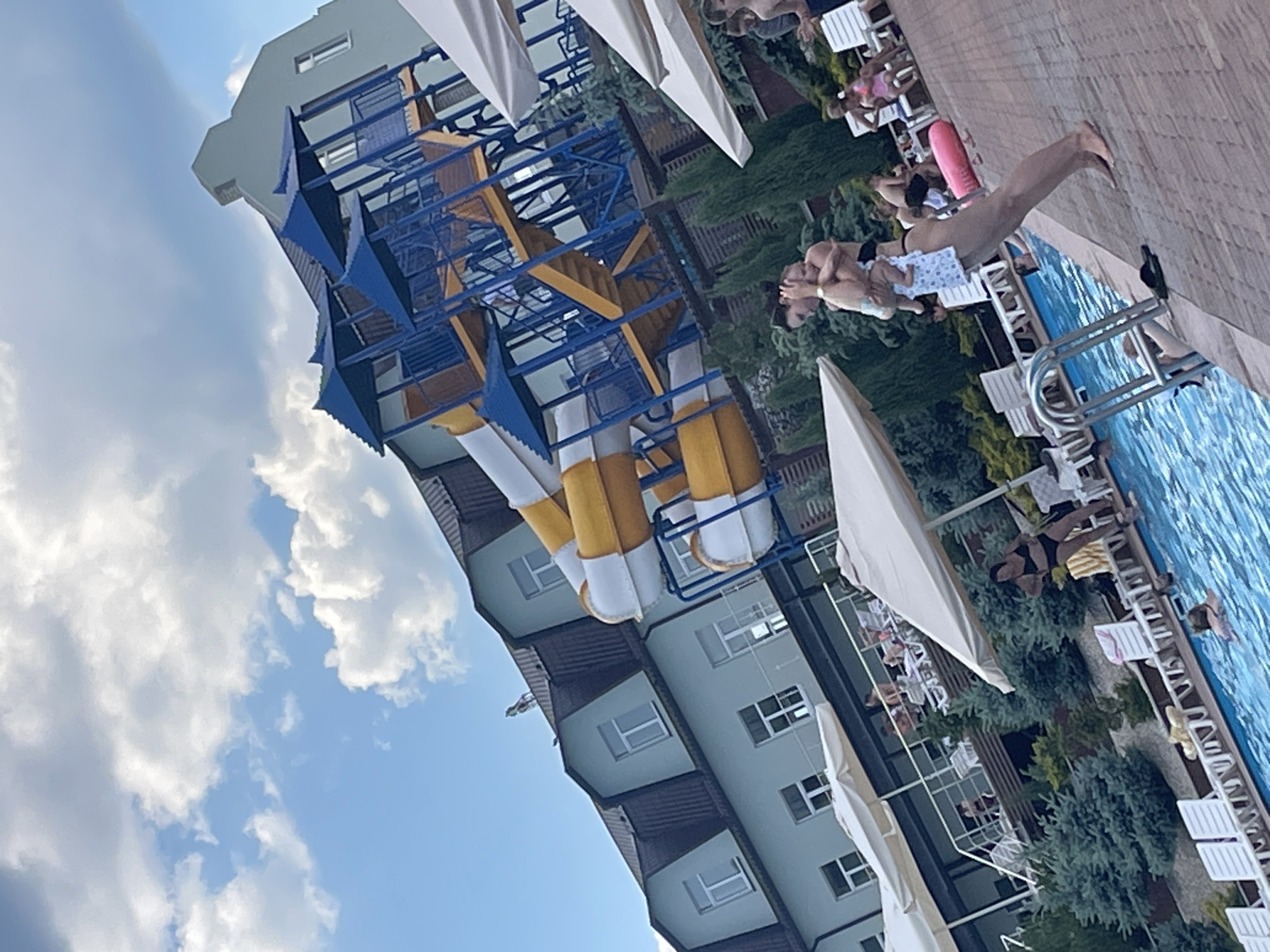 The image depicts a water park, likely located in or near the village of PriShtannoye, Russia, given the context provided. The main structure is a multi-level water slide complex with yellow and white slides, and blue platforms and railings. A light blue building with a dark-pitched roof and dormer windows is situated behind the slides. The foreground features a swimming pool with blue water, a paved deck area with lounge chairs, and several people. In the center of the frame, a woman in a black swimsuit is holding a baby. Other individuals are visible by the pool, some in swimsuits, and some sitting on lounge chairs. Green trees and shrubs are interspersed throughout the scene, and white umbrellas provide shade on the deck. The sky is partly cloudy with white cumulus clouds. The overall impression is a sunny day at a recreational facility.