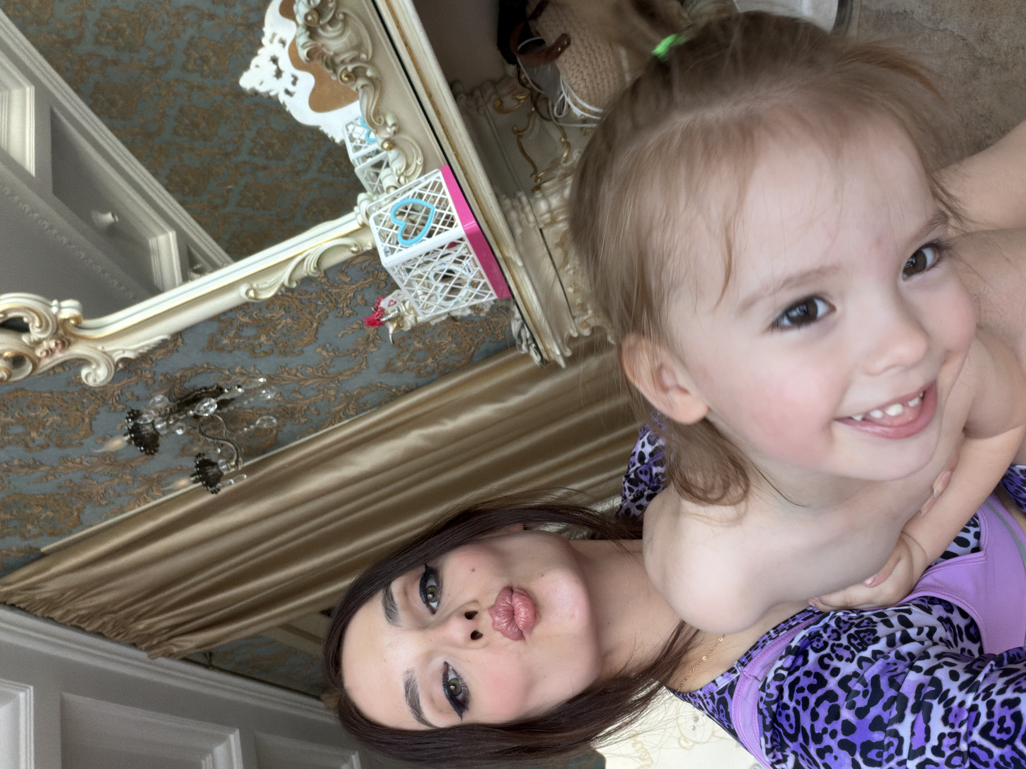 This media file is a selfie featuring two subjects: an adult woman and a young child, likely a mother and daughter. They are posing for the camera in an indoor setting.
The woman in the foreground has dark hair and is wearing makeup, including dramatic eyeliner. She is puckering her lips, giving a playful expression. The child is in the background, closer to the camera, with blonde hair tied up with a green elastic. The child is smiling broadly, showing their teeth. Both individuals are wearing matching purple tops with a black leopard print.
The background suggests a well-decorated interior. A large, ornate mirror with gold trim is visible, reflecting part of the room. The wallpaper has a damask pattern in shades of blue and gold. A chandelier with crystal accents is hanging from the ceiling. Drapes in a golden fabric are also present. The overall impression is of a luxurious or elegantly decorated home.
The lighting appears to be natural, suggesting it's daytime. The emotions conveyed are positive and affectionate, with both subjects displaying smiles or playful expressions. There is no visible text in the image. The location context is Ufa, Russia.