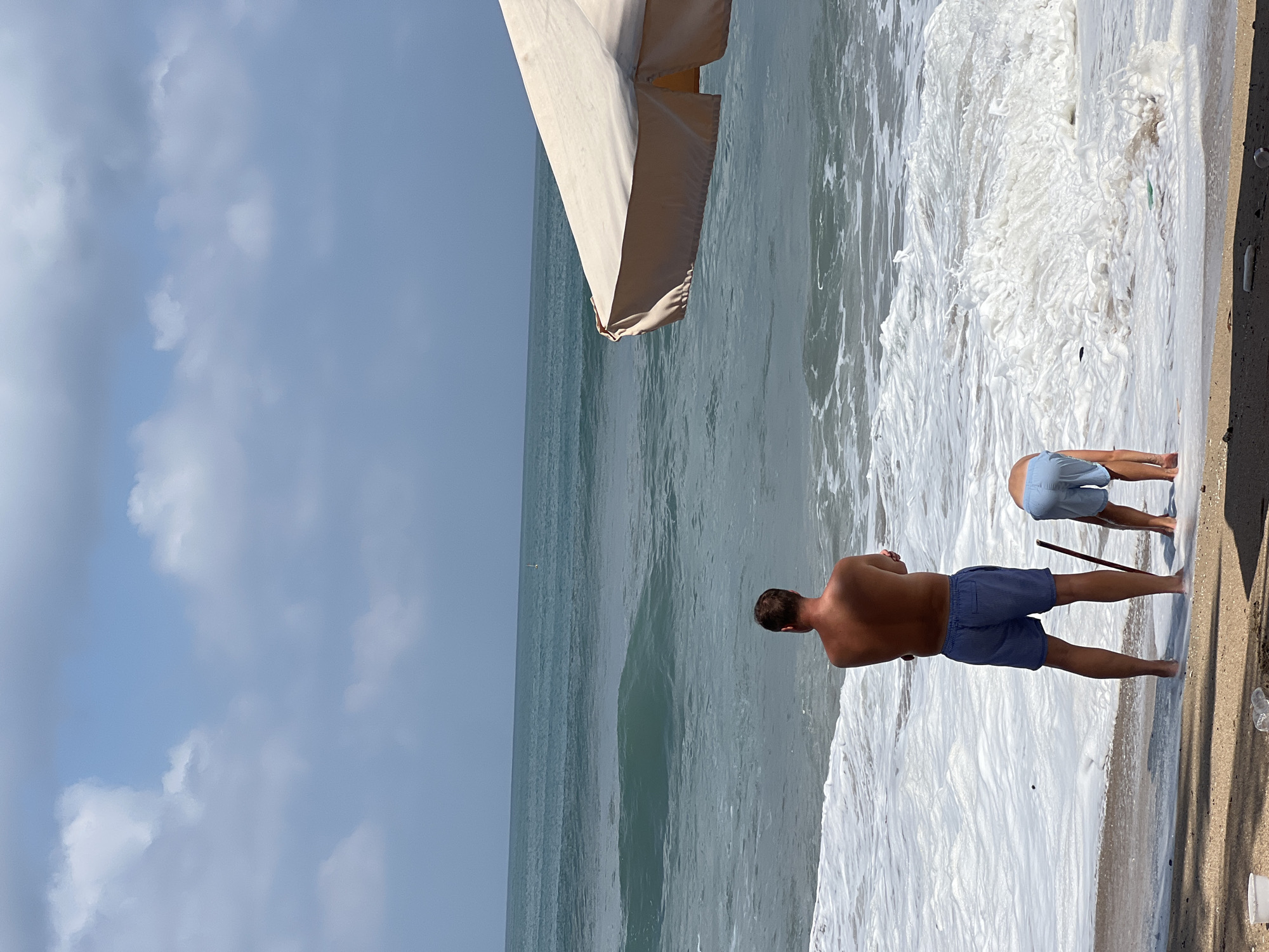 A man and a child are at the beach in Kuta, Indonesia, by the ocean on a cloudy day. The man, facing away from the camera, wears blue shorts and stands in shallow water. The child, also wearing blue shorts, is bent over near the water. A large beach umbrella is partially visible on the right side of the frame. The ocean has moderate waves and a white foamy shore.