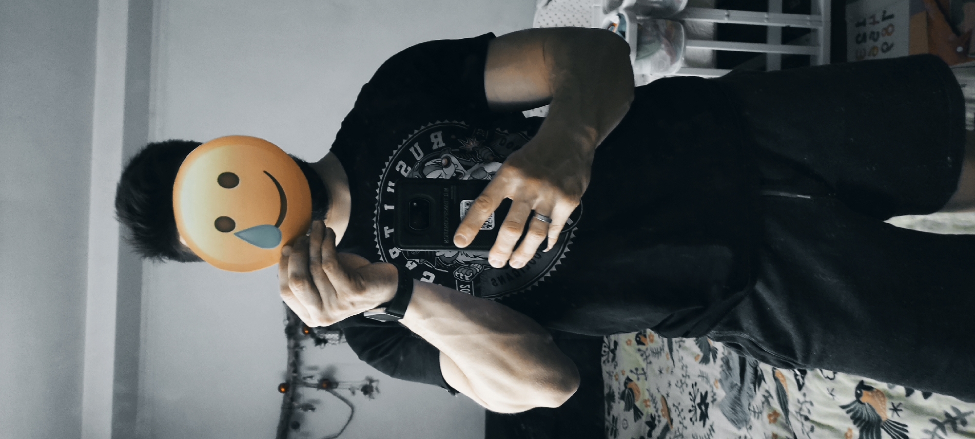 A man stands in a room with light gray walls and floral print bedding. He holds a smartphone in his right hand and a yellow, drooling emoji over his face. He is wearing a black t-shirt, black shorts, and a black smartwatch. His left hand is also visible. The location is a bedroom in Fryazino, Russia.