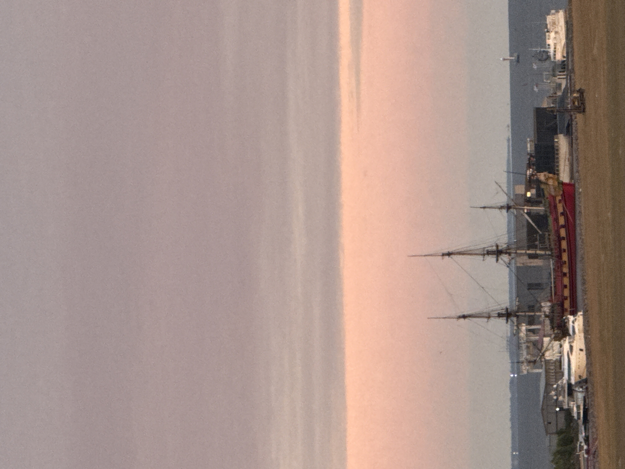 The image depicts a harbor scene at dusk or dawn, with a soft, diffused light illuminating the sky in shades of pink and purple. In the foreground, there is a grassy embankment. Beyond this, a body of water is visible, on which several boats and ships are docked. The most prominent vessel is a large, historical-looking sailing ship with two masts, painted red and yellow. To its left, a modern white yacht is moored. Further in the background, there are industrial buildings and structures, and a distant shoreline. On the water, to the right, a lighthouse is visible, and another white ship is docked. The scene is calm and still, with no visible human activity. It is difficult to ascertain the exact location in Saint Petersburg without further context, as many areas of the city feature harbors and waterfronts.