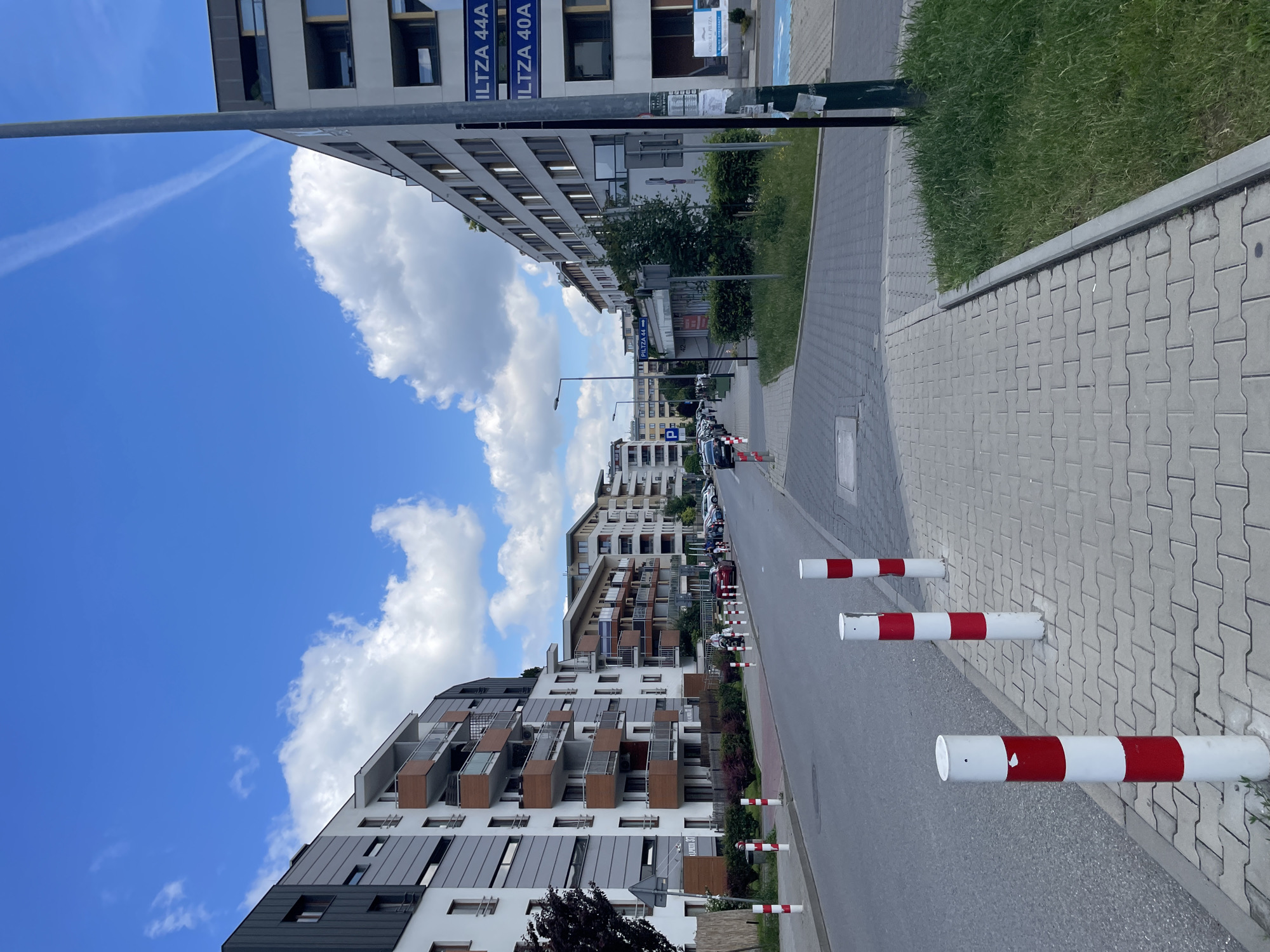 An eye-level, medium shot captures a street scene in a city, possibly a residential or mixed-use area. The perspective is from a sidewalk looking down a road lined with modern apartment buildings on both sides. The sky above is a vibrant blue with scattered white clouds, and a faint contrail stretches across the upper right portion of the frame.
The left side of the street features a multi-story apartment building with a facade of white and grey panels, accented by balconies with wooden elements. Lush green bushes and small trees line the front of the building. Red and white striped bollards are intermittently placed along the sidewalk separating it from the street.
The right side of the street also has a similar apartment building with a light-colored facade and dark-framed windows. A street sign attached to a dark pole indicates "PILIZA 44A" and "PILIZA 40A." Adjacent to the building, there is a patch of green grass and a sidewalk with the same interlocking brick pattern as the left side. Further down the street, more apartment buildings of similar architectural style are visible, and several cars are parked along the curb. People are sparsely visible walking on the sidewalk further down the street. The road surface appears to be asphalt.