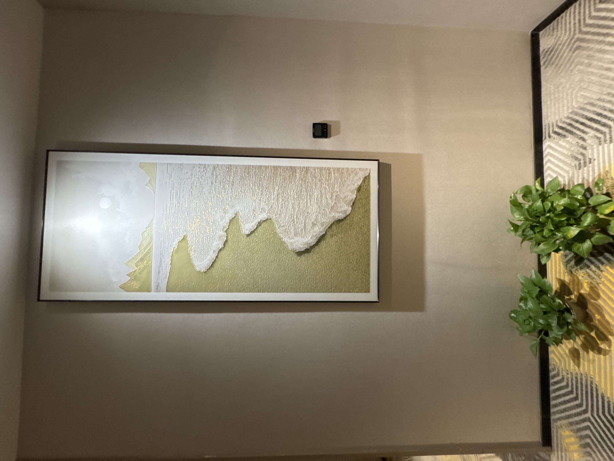In a hallway with tan walls, a tall framed artwork depicts a serene beach scene with golden sand, crashing white waves, and distant mountains under a bright sky. To the right of the artwork, a small black digital device is mounted on the wall. At the bottom of the image, two potted green plants sit on a patterned carpet featuring yellow and grey geometric shapes. The scene is located in Chaoyang District, China.