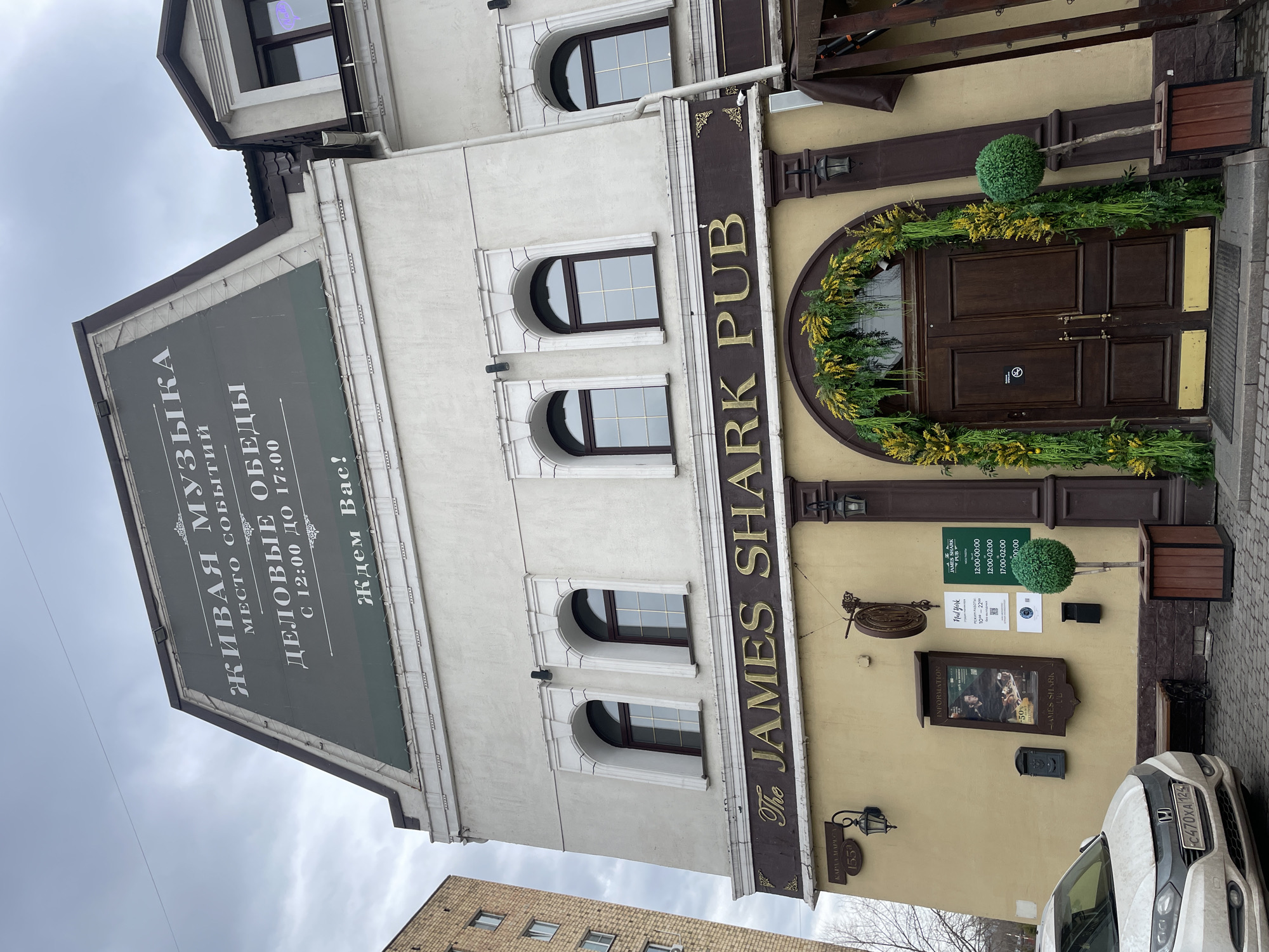 The image shows the exterior of "The James Shark Pub" in Krasnoyarsk, Russia. The pub's signage is visible, as well as a menu board, business hours, and an advertisement for Nail York. The building is multi-story with arched windows and a dark-colored awning/sign with white Russian lettering. The entrance is a wooden double door decorated with greenery. A parked, white Honda CR-V has a license plate C470XA 124. The sky is overcast. Another building is visible in the background, and some bare trees are in the lower part of the image.