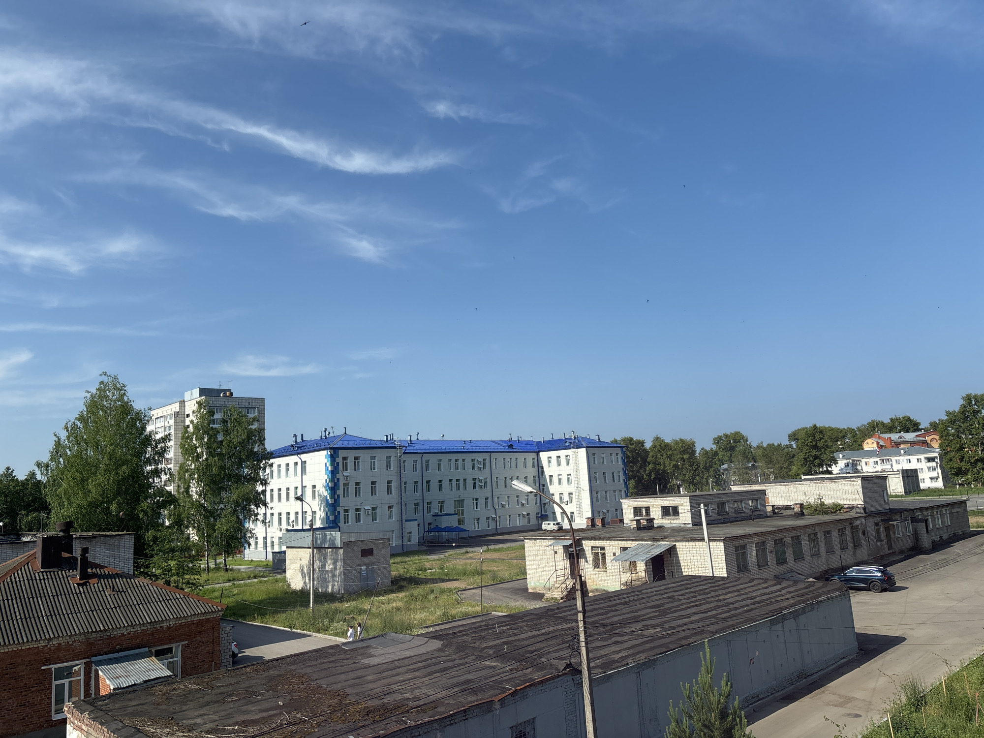 This is a wide, eye-level, outdoor, daylight shot of a semi-urban area with industrial-style buildings and a residential building in Chaikovsky, Russia. The sky is blue with scattered white cirrus clouds and a few dark specks, possibly birds. In the foreground, there are two flat-roofed buildings, one made of red brick with a weathered gray roof and the other with a gray roof and white siding. A person in white is standing near the red brick building. A paved area is visible, with a dark blue SUV parked on it. In the middle ground, there is a long, white, three-story building with a bright blue roof, and to its left, a taller, white apartment building with a darker top. Trees are scattered throughout the scene, and there are smaller, low-rise buildings behind the main structures. The overall impression is one of a functional, somewhat dated, institutional or industrial complex.