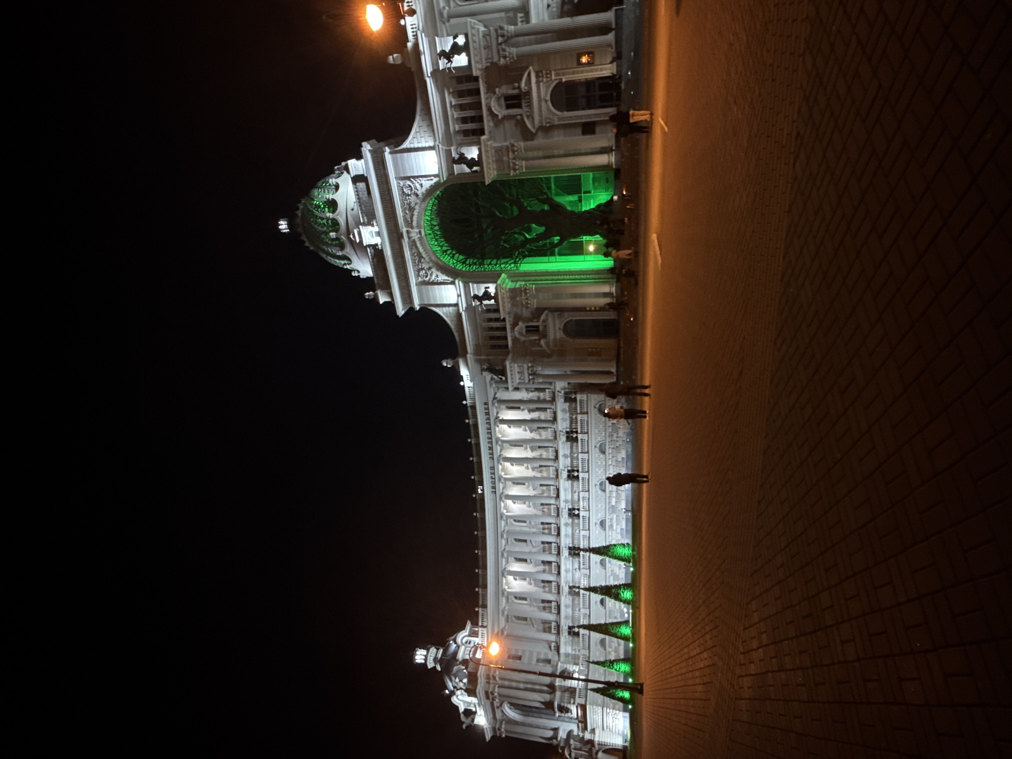 The image captures the Palace of Agriculture in Kazan, Russia, at night. The building, illuminated with white light, features ornate architectural details, including columns, arches, and statues. A prominent green-lit tree is visible within the central archway. Several small, green-lit, cone-shaped decorations line the base of the building. The foreground shows a brick-paved surface, while the background is a dark night sky. A few indistinct figures of people are visible on the brick pavement next to the building.