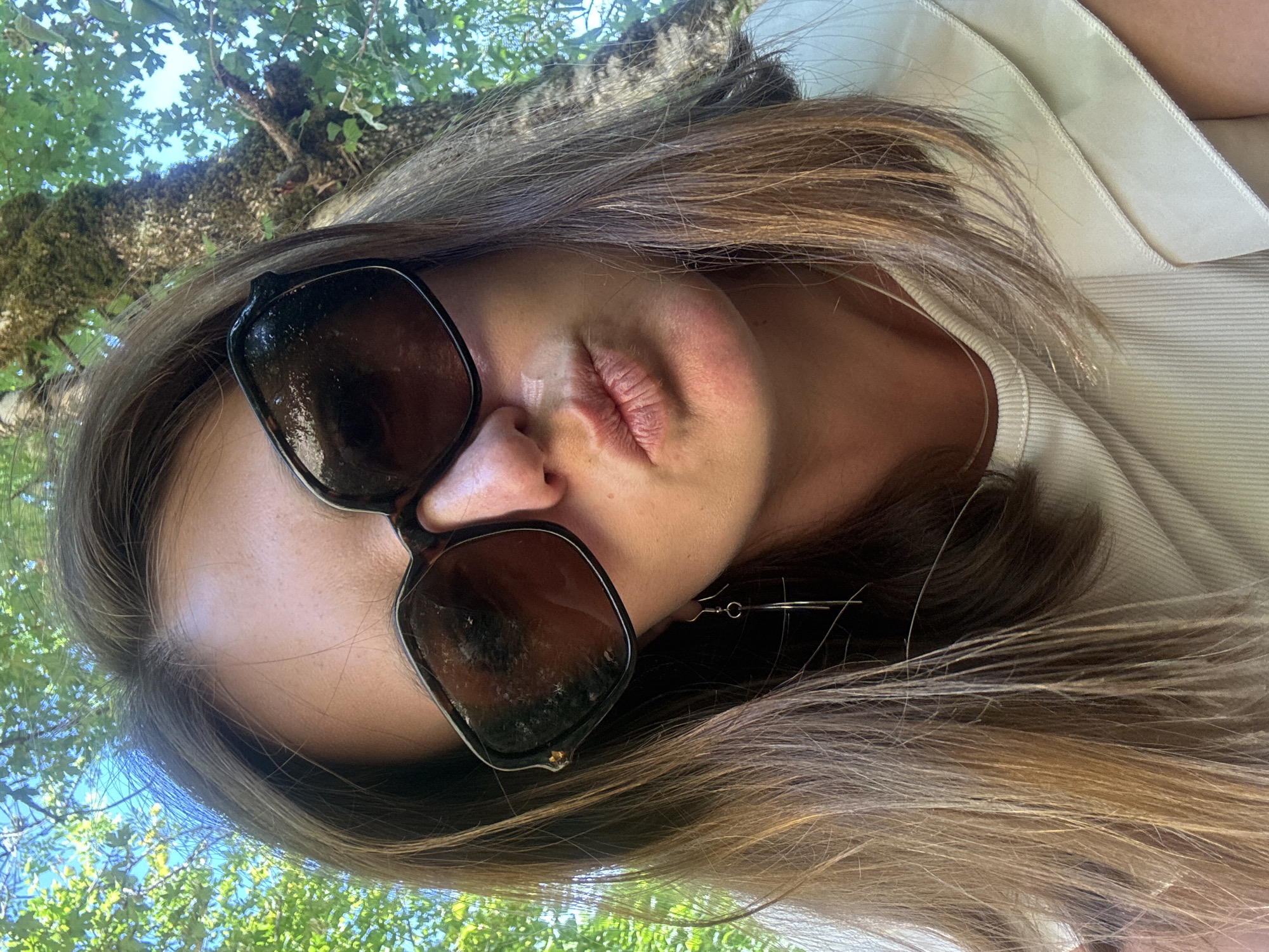 A woman with brown hair is taking a selfie outdoors. She is wearing large, dark sunglasses and a light-colored top with ruffled sleeves. Her lips are pursed. The background shows green foliage and a tree trunk. The image appears to have been taken in Coteaux du Lizon, France.